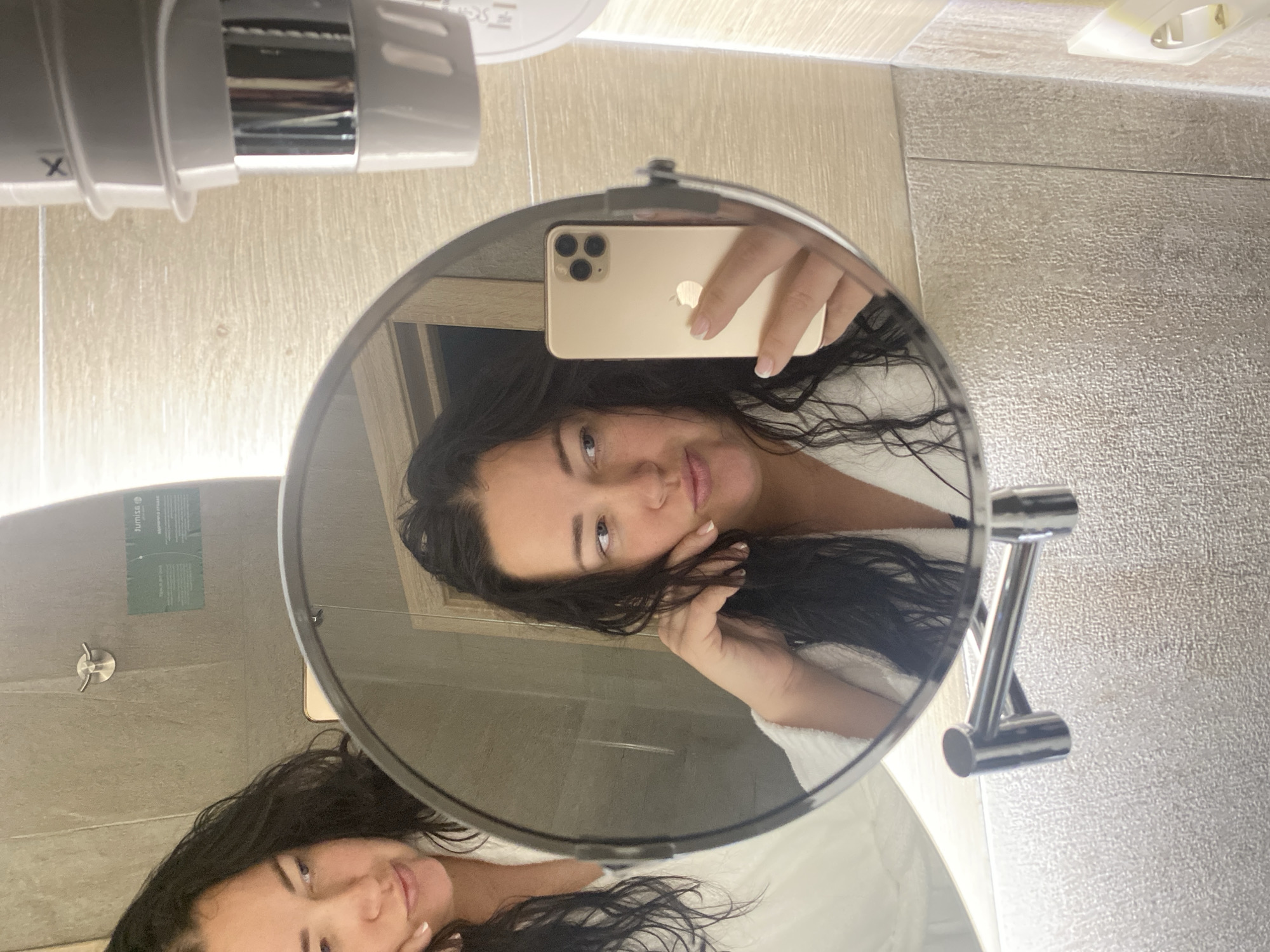 A woman is taking a selfie in a bathroom mirror. She is wearing a white robe and has dark hair. The phone in her hand is gold and appears to be an iPhone. The bathroom has light-colored tiles and a wall-mounted hair dryer. The reflection in the mirror shows a portion of the bathroom, including a shower area with a toiletries dispenser. There is no clear visual evidence to confirm this image was taken in Sochi, Russia.