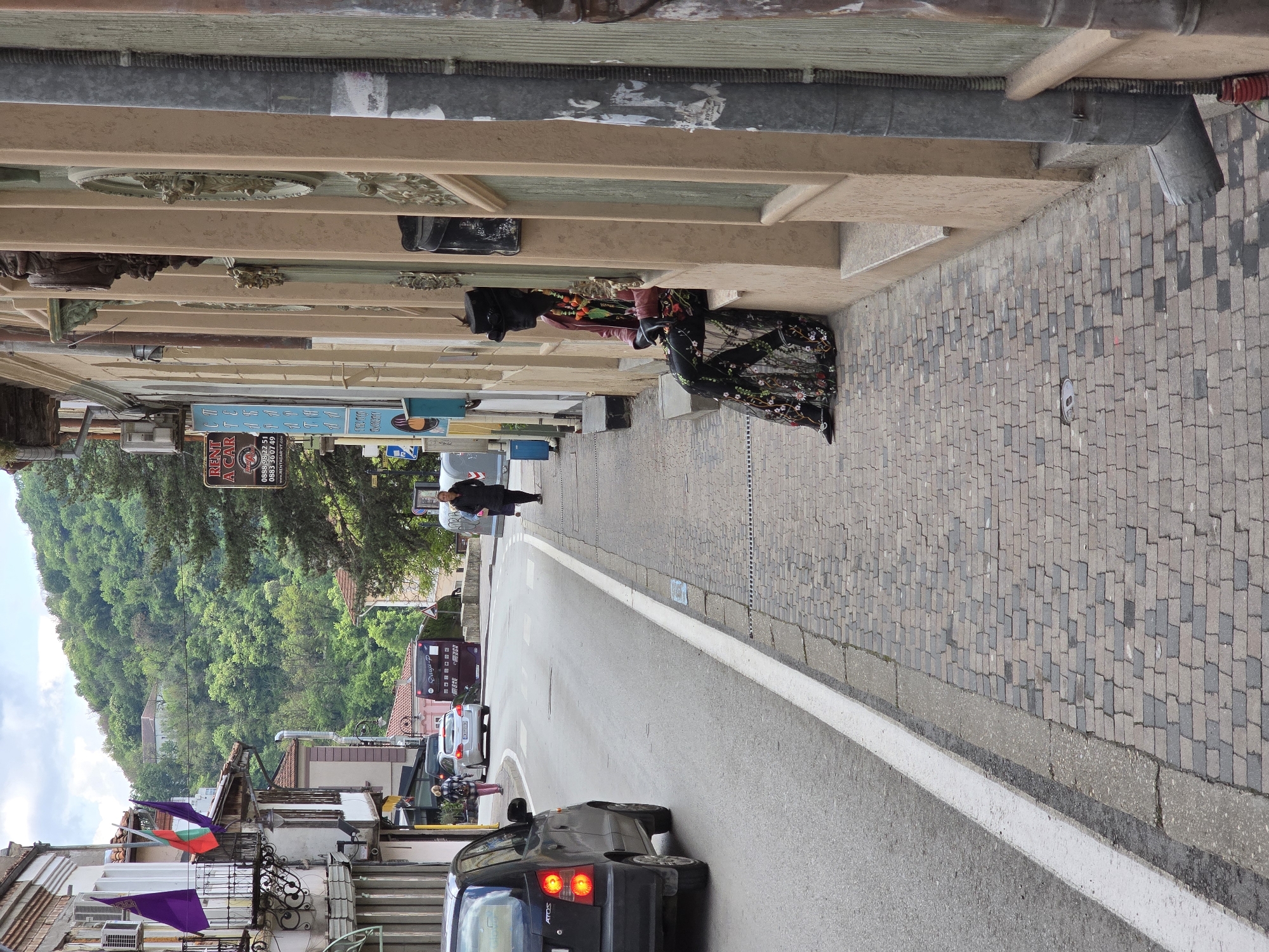 This daytime street scene captures a vibrant urban environment with a lush, green forested hill dominating the background under an overcast or partly cloudy sky. The image is oriented vertically, with the ground level on the right.
On the left side (once rotated correctly), a row of multi-story buildings features ornate architectural details, including decorative reliefs and weathered facades. A grey metal pipe runs along the upper section of these structures. Below, on the paved sidewalk made of grey and dark grey cobblestones, several people are present. Two women are seen near the buildings, one in a dark floral-patterned dress, possibly interacting with goods displayed outdoors. Further down the sidewalk, another individual walks away from the viewer.
The asphalt road in the foreground shows a black Hyundai Atos car with its taillights illuminated, heading away from the viewer. A solid white line delineates the road from the sidewalk. Further down the street, a lighter-colored car and a dark bus or large van are visible. From a balcony on one of the buildings, purple and green/red flags are draped.
Visible text on signs includes "RENT A CAR," phone numbers "0888 98 22 51" and "0883 36 0749," and a website "www.RENTACAR-VT.COM." Another sign displays "Олци-Р." The "ATOS" brand name is visible on the rear of the black car.