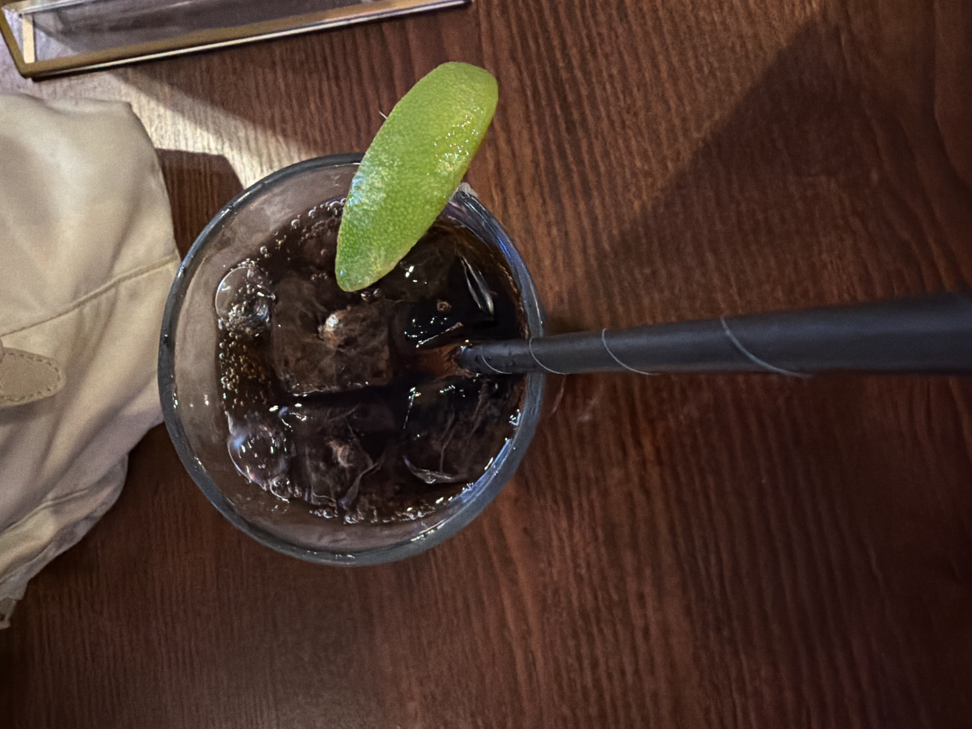 An overhead shot captures a glass of dark, carbonated beverage, likely a soda or cocktail, resting on a wooden table. The glass contains several ice cubes, and small bubbles are visible on the liquid's surface and around the ice. A vibrant green lime wedge is perched on the rim of the glass, adding a fresh accent. A black drinking straw extends from the drink towards the right side of the frame. In the upper left portion of the image, a light-colored, possibly beige, piece of cloth or napkin is visible, along with a sliver of a metallic or framed object. The table surface is made of dark wood with a distinct grain, and the lighting suggests an indoor setting, such as a restaurant or bar in London, United Kingdom. No people are visible, and there is no discernible text. The image presents a static scene focused on the drink, possibly awaiting consumption.
