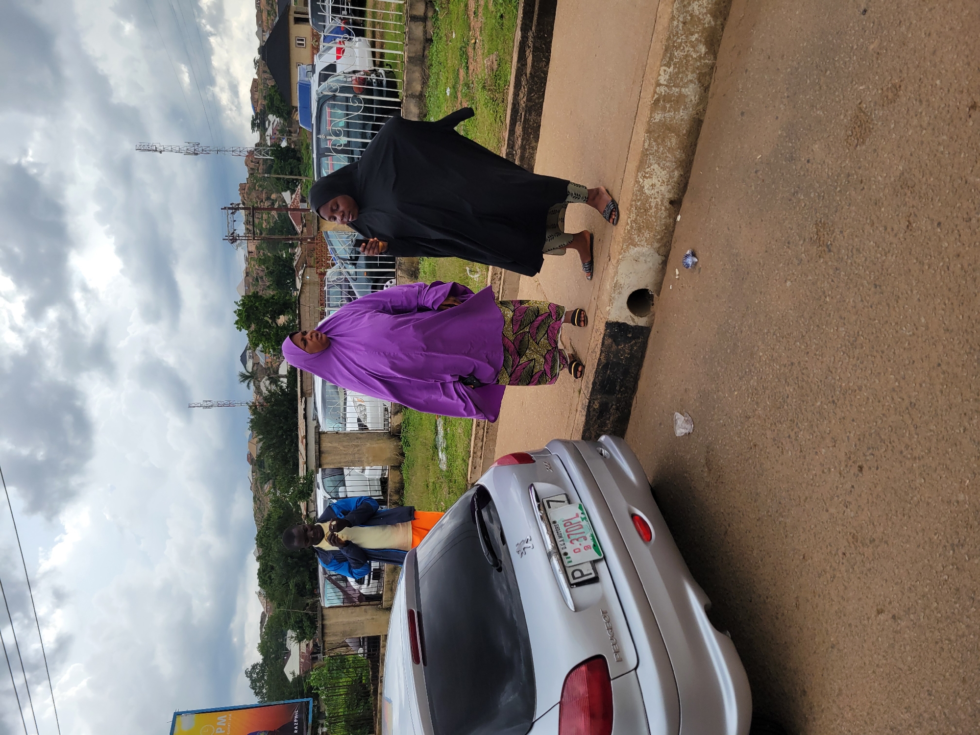 In Jos, Nigeria, under a cloudy sky, a silver Peugeot 206 is parked on the right side of a paved road. A man in a blue jacket, yellow shirt, and orange trousers stands on the sidewalk, looking down at something he is holding. To his right, a woman in a bright purple hijab and patterned skirt stands, facing left. Next to her, a woman in a black hijab and patterned pants holds a phone to her ear. The sidewalk has a concrete edge with a drainpipe. Behind them, a metal fence separates the sidewalk from an area with parked cars and buildings. A billboard is visible on the far left.