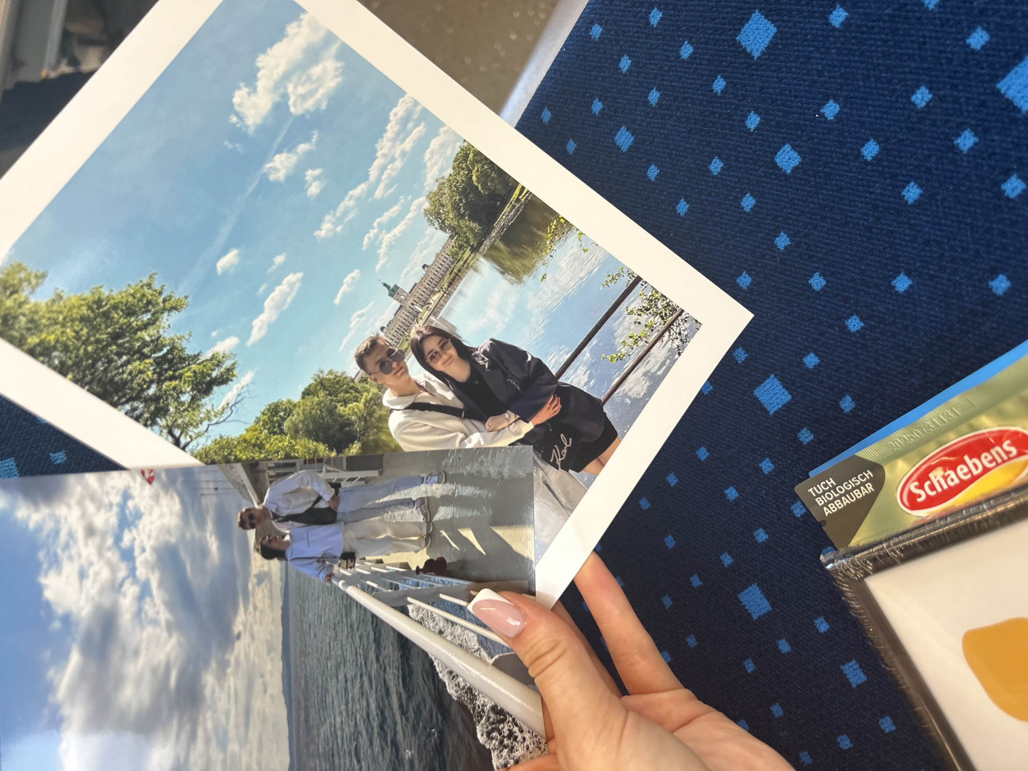 A close-up shot captures a person's hand holding two printed photographs against a dark blue patterned background, likely upholstery in a vehicle.
The prominent photo, held by a hand with a neat French manicure, features a young couple outdoors on a bright, sunny day. The man, wearing sunglasses and a light top, has his arm around the woman, who is also wearing sunglasses and a dark jacket, possibly with a "KARL" logo visible. They stand beside a metal railing, overlooking a calm body of water, with a grand, ornate building (resembling a palace or castle) and green trees in the background under a blue sky with white clouds.
Behind this main photo, a second, partially visible print shows two other individuals standing on a boat deck, with water and distant land or hills under a bright, cloudy sky.
To the right, on the blue patterned background, a product package from the brand "Schaebens" is partially visible. The text "TUCH BIOLOGISCH ABBAUBAR" (German for "CLOTH BIOLOGICALLY DEGRADABLE") can be read on the package. The patterned surface consists of small, light blue diamond shapes. The overall impression is of someone reviewing cherished memories, perhaps during travel.