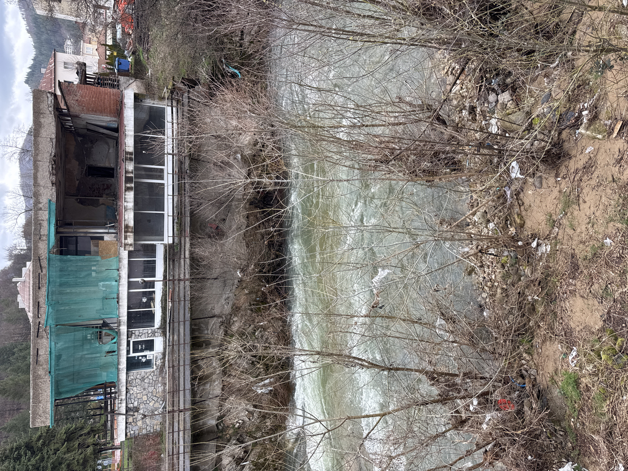The scene presents a river cutting through a residential area in Rudozem, Bulgaria. The river appears fast-flowing and murky, likely carrying silt or debris. The riverbanks are strewn with trash, including plastic bags, bottles, and other discarded items, suggesting a lack of proper waste management. Along the river's edge, a building stands, partially constructed or under renovation, with exposed brick and areas covered in green netting. The architectural style is modern, with large windows and a multi-level structure. The surrounding vegetation consists primarily of bare trees and sparse ground cover, indicative of late fall or early spring. The sky is overcast, casting a diffused light across the scene. The overall impression is one of neglect and environmental degradation within an urban environment.