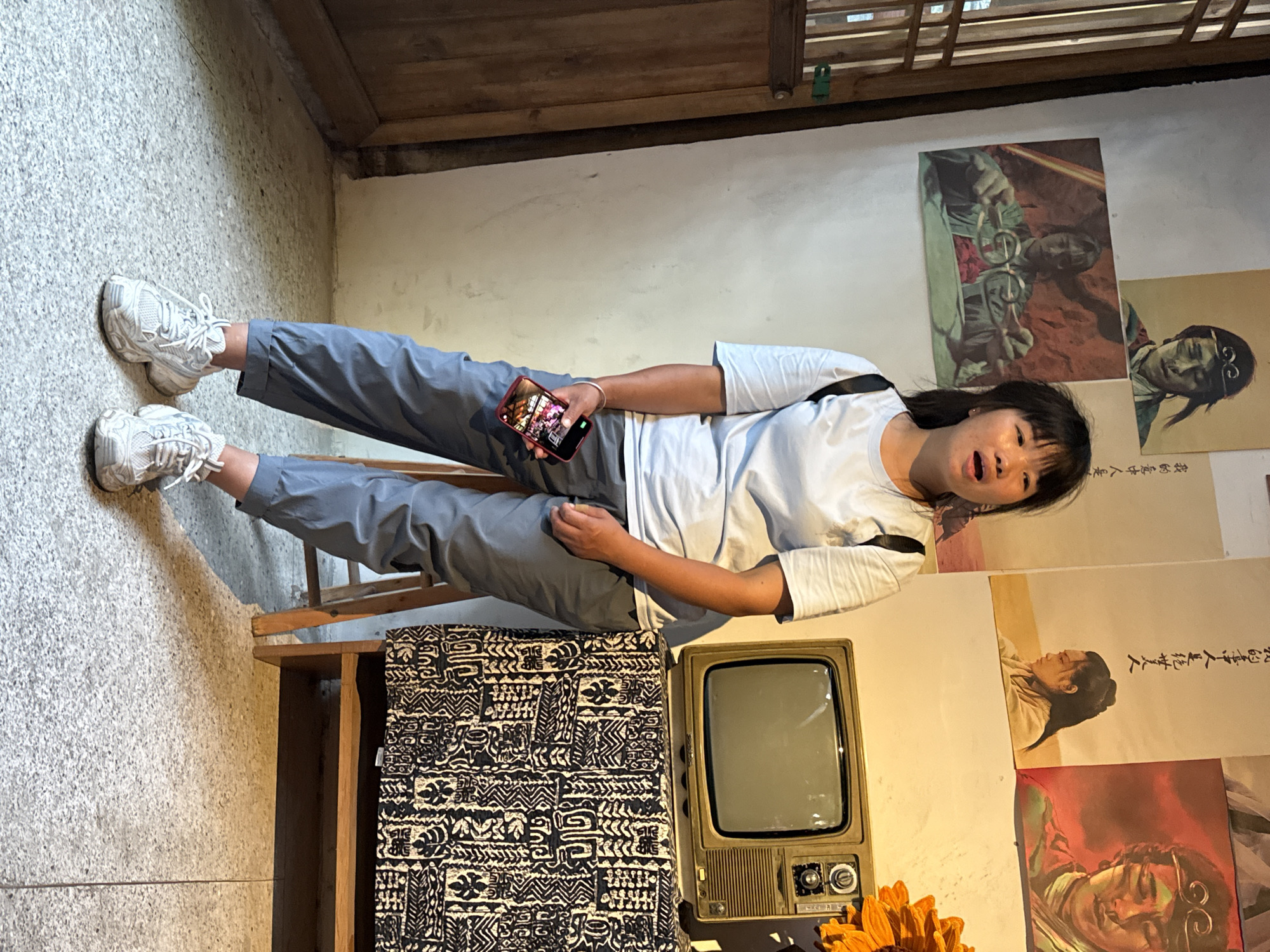 The image shows a woman sitting on a chair, holding a mobile phone in her hand, and looking upwards. She is wearing a white T-shirt and gray pants, and white sneakers. Behind her are various pictures and posters, and a vintage television set is placed on a black and white patterned fabric-covered stand. The setting appears to be indoors, possibly a shop or a themed area in Beibei, China.
