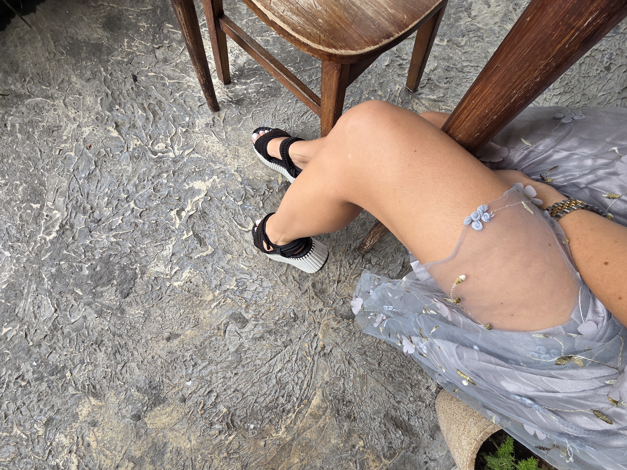 An overhead shot shows a person sitting on a wooden chair, with their legs crossed. The person is wearing a sheer, light blue dress with floral embellishments and black and white sandals. Their right leg is bent at the knee, and their left leg is extended. The ground beneath them is a textured, grey surface with a rough, uneven pattern. A portion of a wooden chair leg is visible in the lower right corner of the frame. The setting appears to be outdoors, possibly a patio or garden area. The image does not contain any elements that would allow for specific identification of the location within North Kuta, Indonesia.