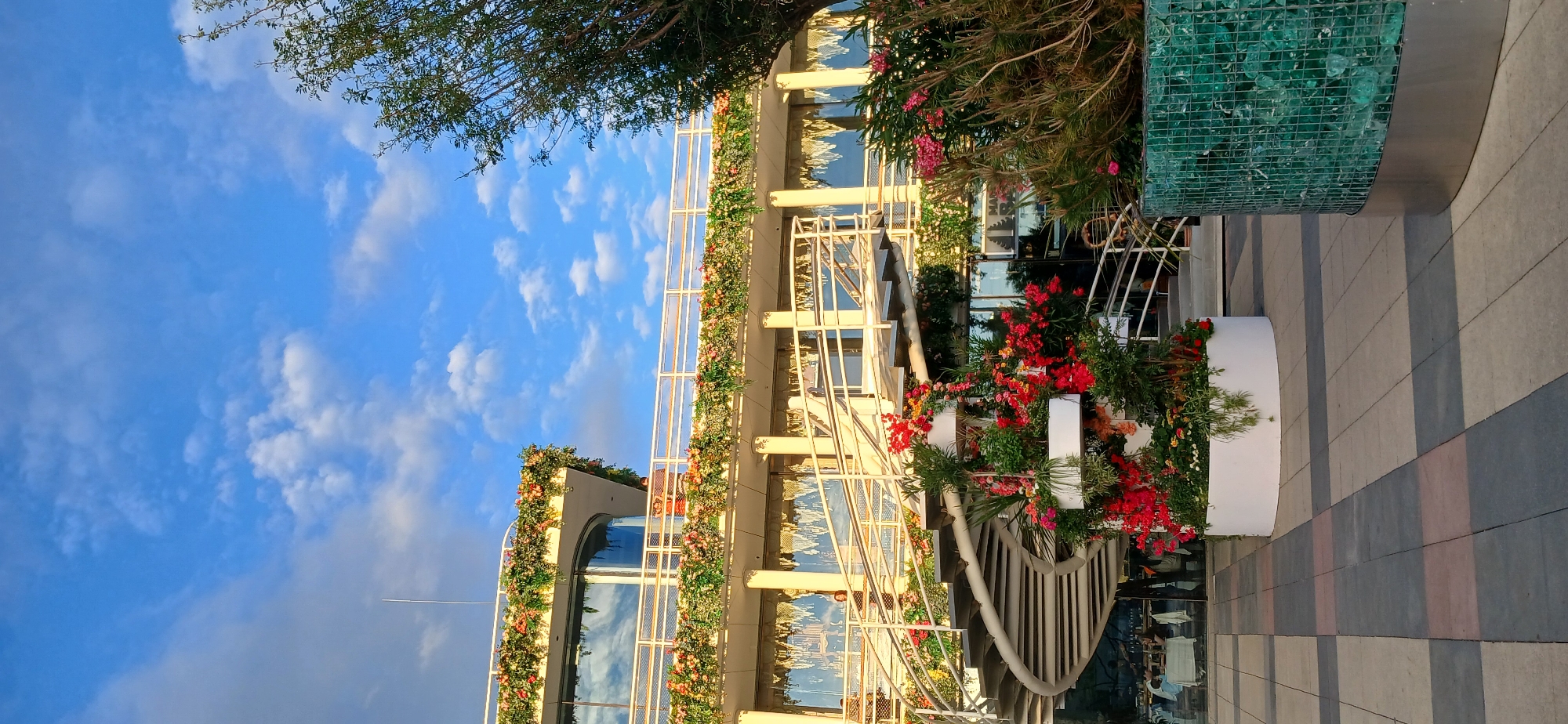 A spiral staircase adorned with abundant red and pink flowers winds upwards outside a multi-story building. The building has large windows and is decorated with floral garlands along its balconies. The sky above is blue with scattered white clouds, and a tree branches into the frame from the right. The ground is paved with a checkered pattern of gray, pink, and black tiles.