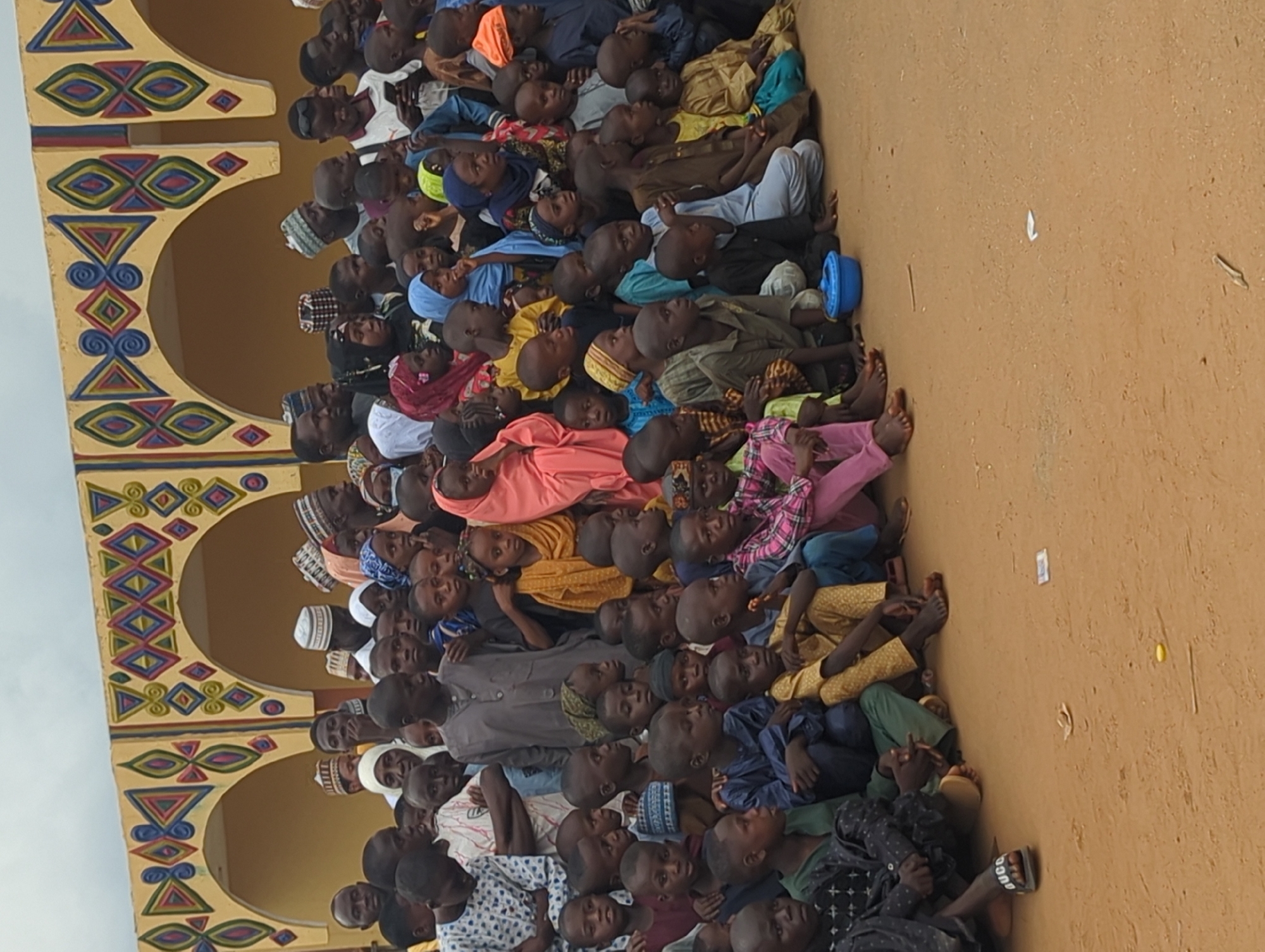 A large group of people, mostly children and some adults, are sitting on the ground beside a building with arches and colorful geometric patterns. The ground is dirt or sand. The scene is likely outdoors in Kafanchan A, Nigeria. The people are facing in the same direction, many looking up. Some wear traditional clothing or head coverings.