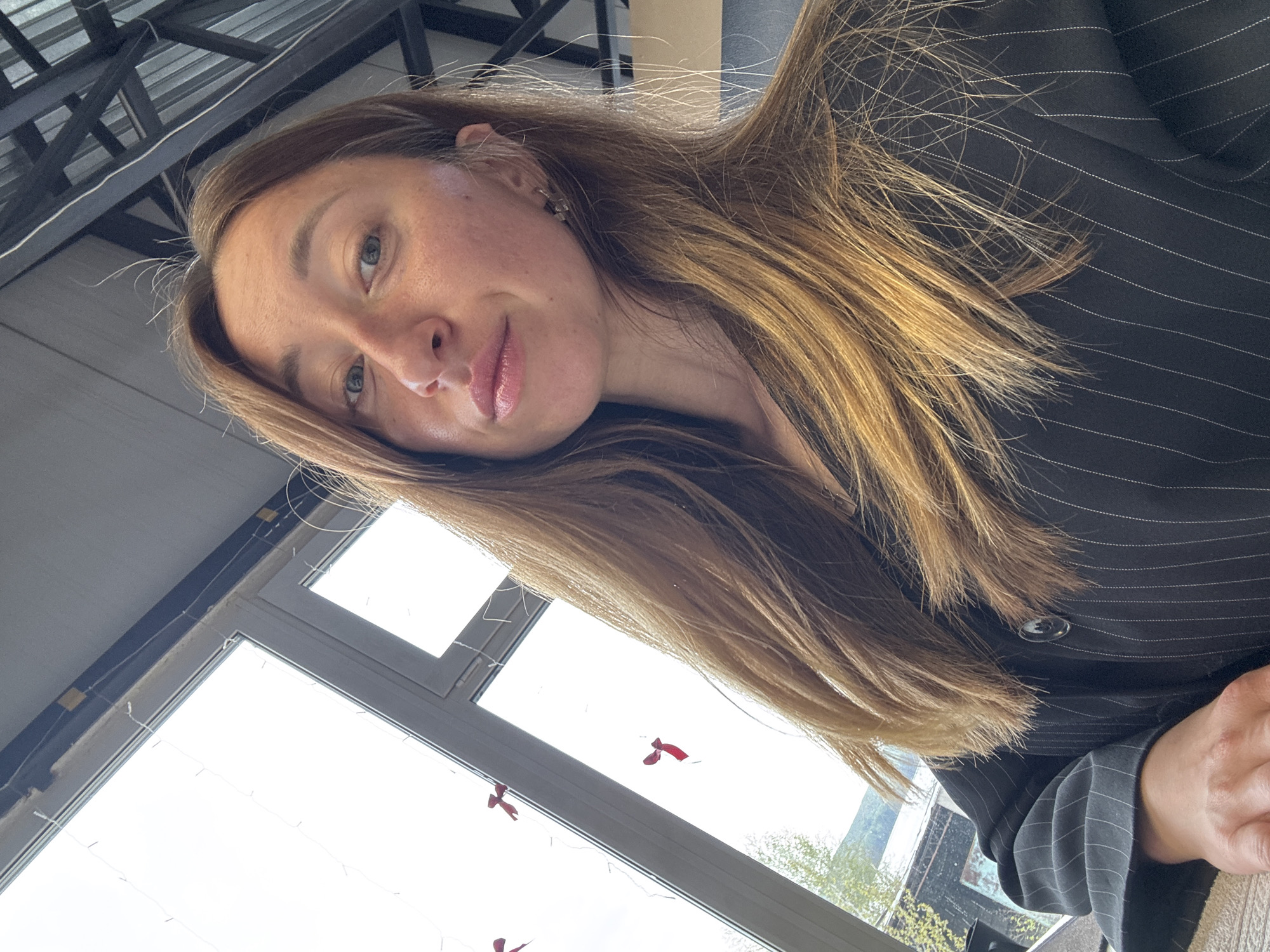 A young woman with long, light brown hair is photographed indoors. She has fair skin, light eyes, and her hair is styled to the side. She wears a dark pinstripe blazer. In the background, there's a window with daylight shining through and a ceiling structure above. The location is potentially Chaikovsky, Russia, but there is no definitive information in the provided image.