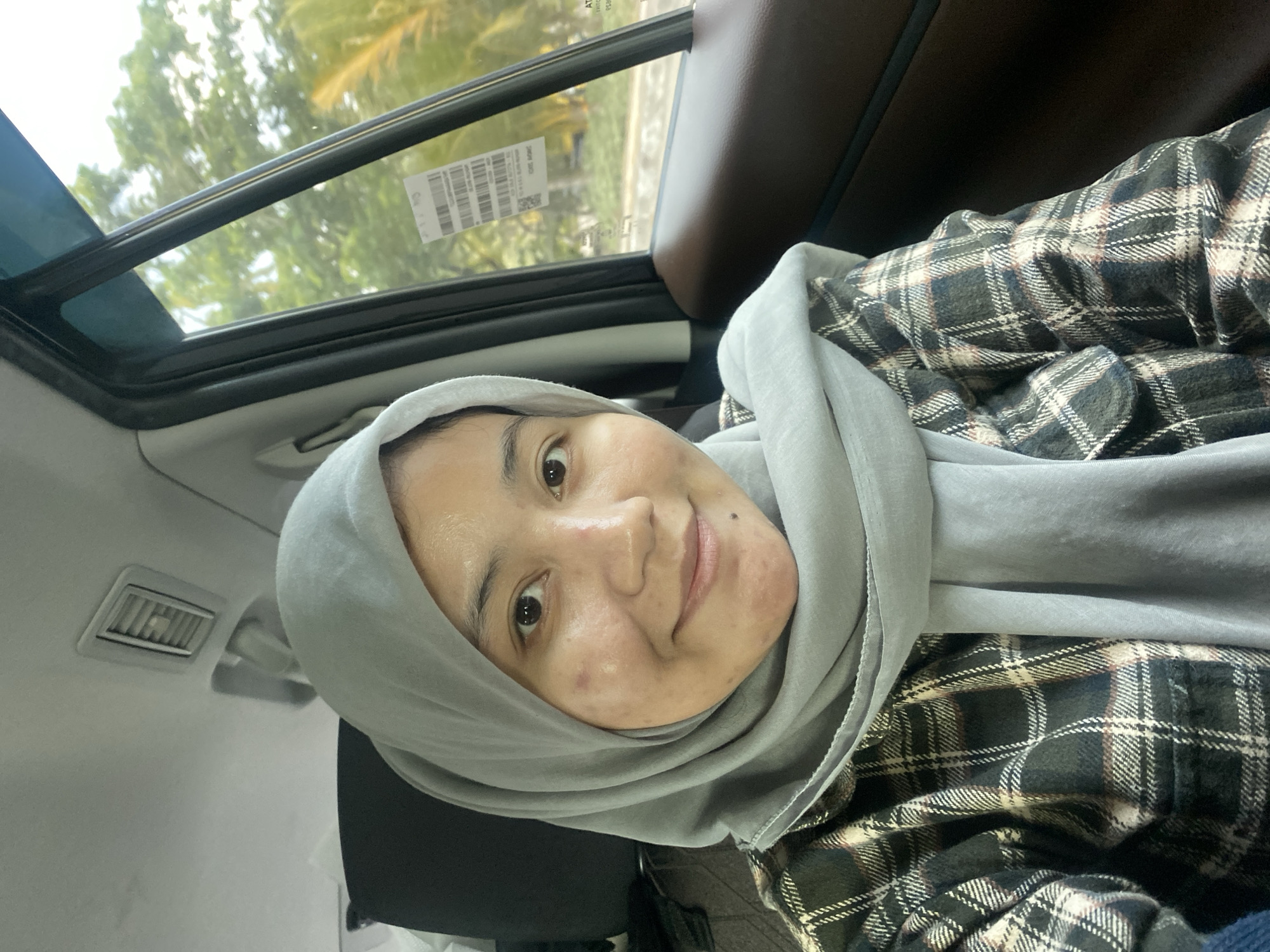 The image is a selfie taken from inside a car in Banjarbaru, Indonesia. A woman wearing a light gray hijab and a plaid shirt is visible. She is looking at the camera and smiling. The interior of the car, including a vent on the ceiling and a window with greenery outside, is also visible. A sticker is affixed to the car window.