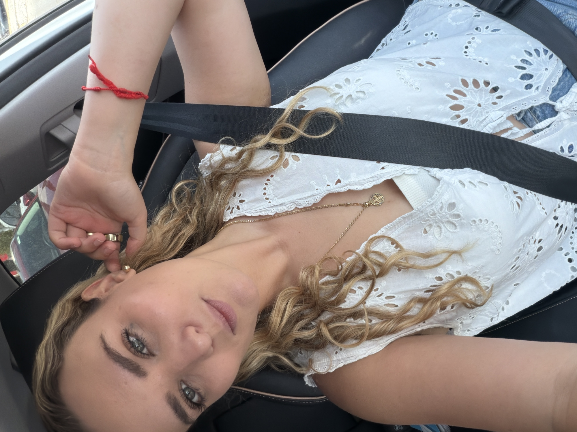 A woman is sitting in the driver's seat of a car, wearing a white eyelet-embroidered top and a seatbelt. She has blond curly hair, fair skin, and is wearing a gold necklace. A red string bracelet is on her left wrist, and she is wearing a gold earring and ring. The location is likely in Puebla, Mexico, based on the background seen through the car window.