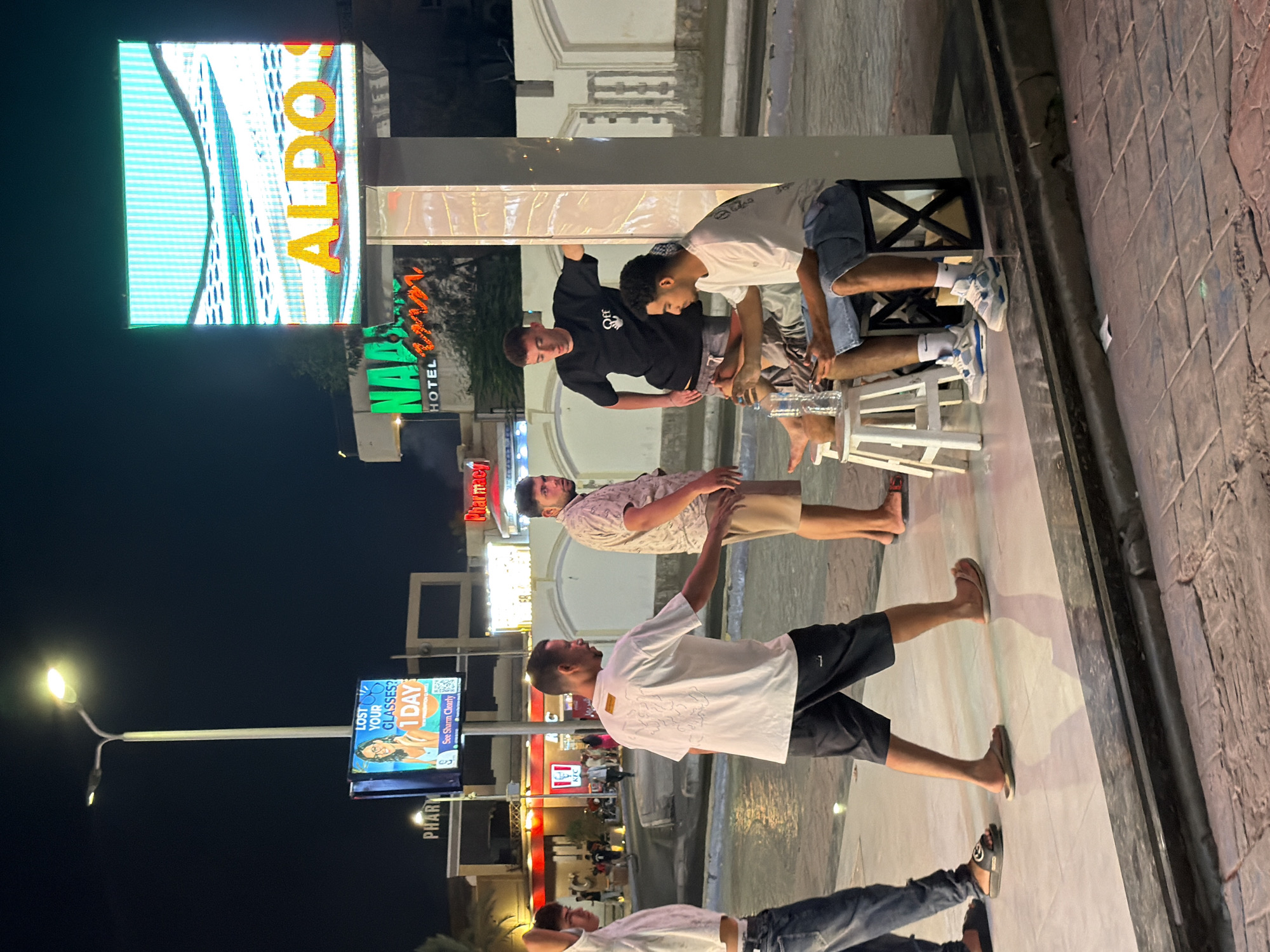 The image depicts an outdoor nighttime scene in Sharm El Sheikh, Egypt, specifically on a promenade likely within a commercial area given the signage. In the foreground, four young men are visible. Two are standing, one wearing a white t-shirt with black shorts and flip-flops, the other in a black t-shirt and grey shorts. The first man is walking away from the camera, while the second is looking to his right. Two other men are seated. One, wearing a white t-shirt and blue shorts, is looking down at a phone and holding a bottle of water. The other, wearing a light-colored patterned short-sleeved shirt and beige shorts, is partially obscured, looking towards the camera.
Behind the group, several elements indicate a bustling urban environment. A tall street lamp with a bright light illuminates the scene. A digital billboard displays the text "ALDO HOTEL" in large, illuminated letters, suggesting a commercial establishment. Another smaller screen shows an advertisement for an optician, featuring the text "LOST YOUR GLASSES? 1 DAY" and an image of a woman wearing glasses. Further back, a KFC restaurant sign is visible, along with other illuminated shopfronts and buildings. The ground is paved with tiles and cobblestones, and a reflective surface, possibly water or a wet pavement, is present in the mid-ground. The overall atmosphere is one of an active evening in a tourist or commercial district.