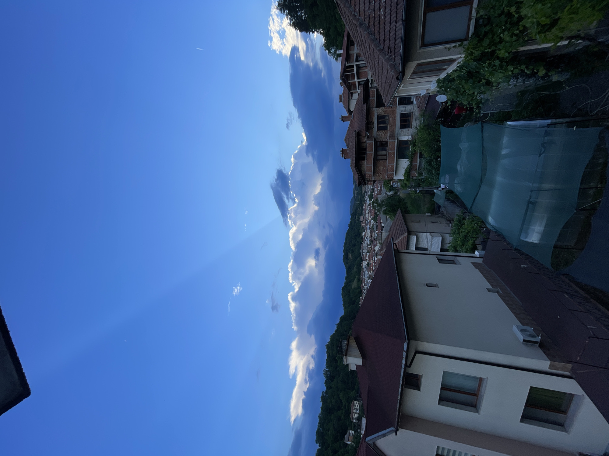 The image depicts a view of the city of Zlatograd, Bulgaria, from an elevated vantage point, likely a balcony or rooftop, as indicated by the presence of a dark, textured overhang in the upper left corner. The sky is a prominent feature, displaying a vibrant blue with dramatic cloud formations. Sunbeams, or crepuscular rays, are visible, piercing through the clouds from the right side of the frame, creating a sense of depth and atmospheric drama.
The landscape below is characterized by a dense urban environment interspersed with green hills. Numerous houses with terracotta tiled roofs are visible, clustered together on the slopes. The architectural style suggests a traditional Balkan town. In the foreground, the roofs of several houses are clearly visible. One house in the immediate foreground on the left is painted a light color and has a dark red roof with a prominent chimney. To its right, another house features a brick facade with multiple balconies and large windows. A green mesh-like structure, possibly a covered patio or greenhouse, is spread out between the houses in the mid-ground. Lush green trees and foliage are present, particularly on the surrounding hillsides and climbing up some of the structures, adding to the natural beauty of the setting.
The time of day appears to be late afternoon or early evening, given the quality of the light and the intensity of the blue in the sky. There are no people or active events depicted in the image. The scene is static and serene, focusing on the interplay of natural light and the urban landscape of Zlatograd.