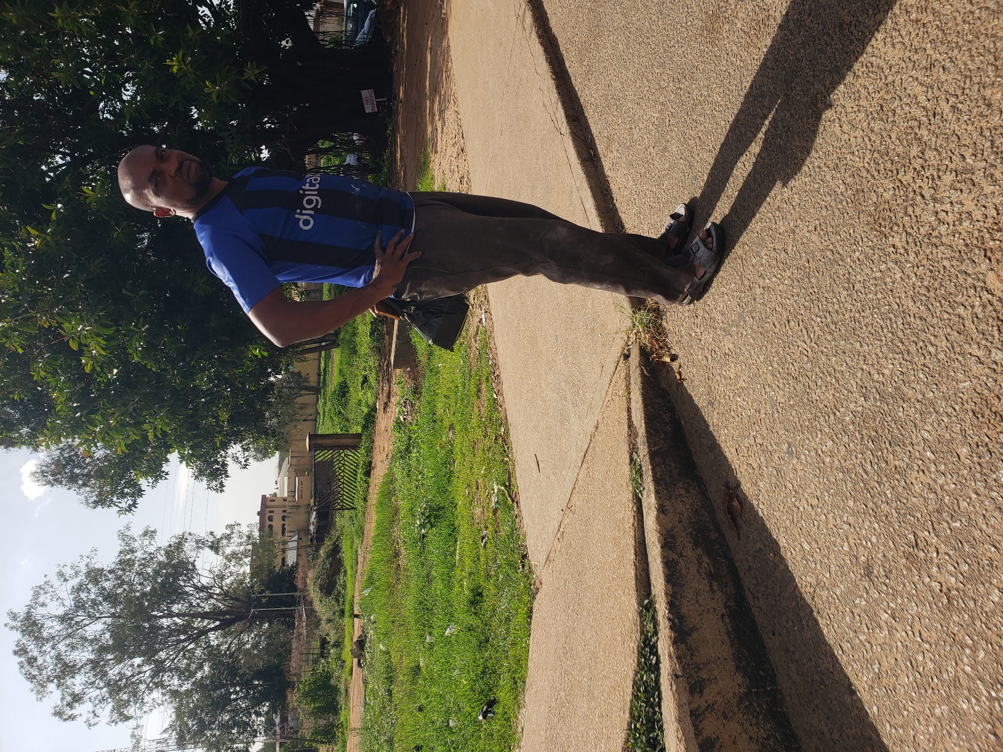 In Gura Top, Nigeria, a bald, dark-skinned man with a goatee stands on a concrete sidewalk. He is wearing a blue and black striped soccer jersey with "digital" written on it in white, and dark brown pants. A black bag hangs from his left shoulder. He has his hands on his hips and is looking to the right. Behind him, a large tree with dark green leaves stands near a dirt road. In the background, there is a building and some vegetation. The sun casts long shadows across the sidewalk.