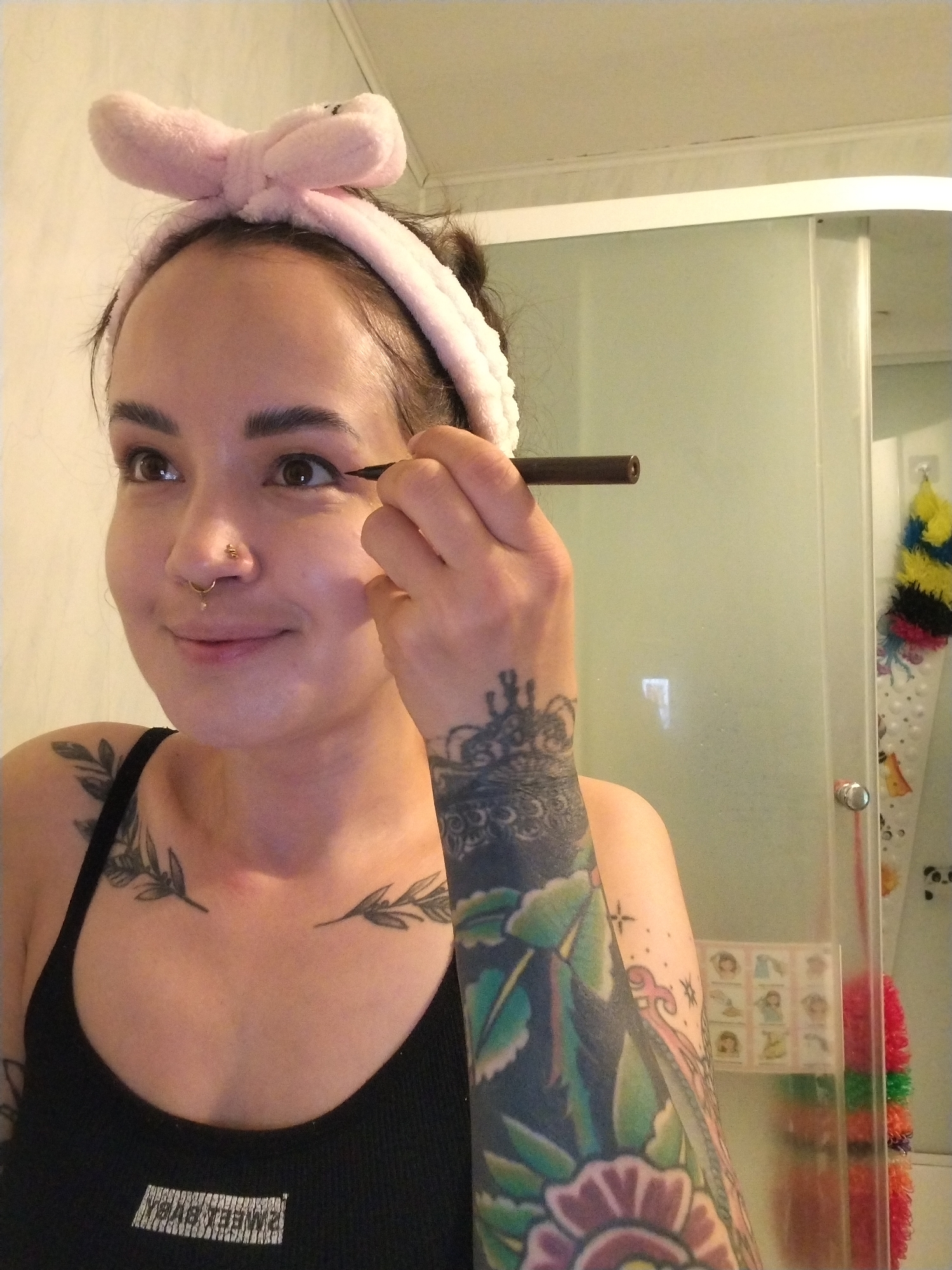 A woman is captured in an indoor setting, likely a bathroom in Noviy, Russia, applying eyeliner to her right eye. She is looking slightly upwards and to her right, presumably into a mirror which is not fully visible. Her dark hair is pulled back, held by a soft pink headband shaped like a bow. She has a gentle, pleasant smile on her face, indicating a sense of calm or contentment.
The woman wears a black tank top, on which white, mirrored text, possibly "SWEET BABY," is partially visible across her chest. She has several facial piercings, including a gold hoop with a small dangling bead in her septum and a stud on the side of her right nostril. Her arms and chest are extensively tattooed; a dark, ornate crown-like design covers her right forearm, while a vibrant, colorful sleeve featuring floral and leaf motifs adorns her left arm. Delicate leafy patterns also extend across her collarbones.
The background shows light-colored walls and the partial view of a shower stall with translucent glass. A multi-colored bath puff in shades of yellow, red, and blue hangs inside the shower enclosure. Additionally, a grid of small, framed pictures or cards can be seen affixed to the wall next to the shower. The scene is illuminated by artificial indoor lighting.