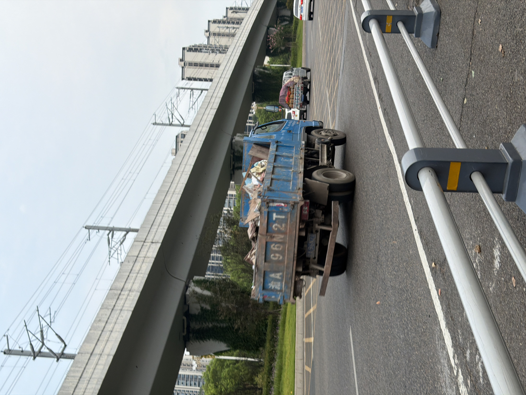 The image depicts a street scene in Jiangjin, China, with a blue dump truck carrying debris driving on a road under an elevated structure. The elevated structure appears to be a highway or a railway line, with overhead power lines visible. Tall buildings line the background, indicating an urban or suburban setting. Other vehicles, including a white car and a van, are also present on the road. The weather appears overcast. The truck has a license plate that reads "A 96N2T".
