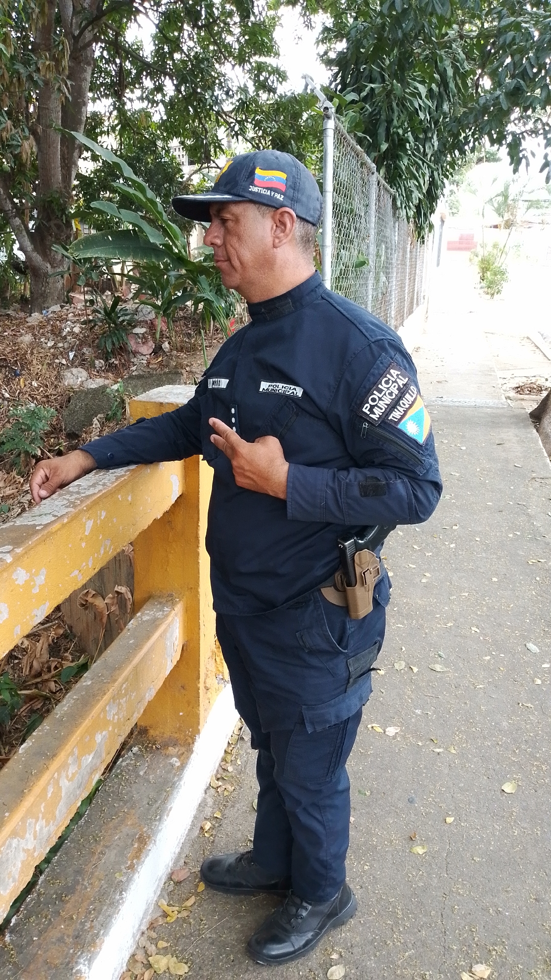 A man in a dark blue police uniform stands outdoors, likely in Tinaquillo, Venezuela, based on the patch on his sleeve. He wears a baseball cap with the Venezuelan flag and the words "JUSTICIA Y PAZ." Patches on his upper arms read "POLICIA MUNICIPAL" and "POLICIA MUNICIPAL TINAQUILLO." A holstered handgun is visible on his right hip. He is leaning against a yellow and white concrete barrier with foliage visible behind it. He's making a peace sign with his left hand. A chain-link fence runs behind him, and trees are in the background. The ground is paved.