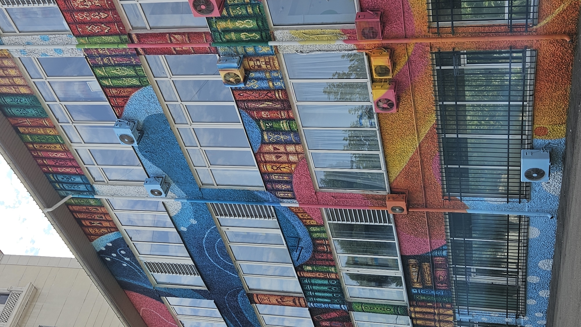 The image shows a building facade decorated with a colorful mural depicting a bookshelf. The mural features rows of intricately painted books in various colors and designs, running horizontally across the building's exterior. The building itself has multiple stories with large windows, some of which are covered with white curtains. Several outdoor air conditioning units are mounted on the walls, some painted in bright colors to match the mural. A black metal fence is installed in front of the lower-level windows. The overall impression is a vibrant and artistic transformation of an ordinary building. This mural is located on the facade of the Almaty Central City Children's Library, a prominent cultural institution in Almaty, Kazakhstan.