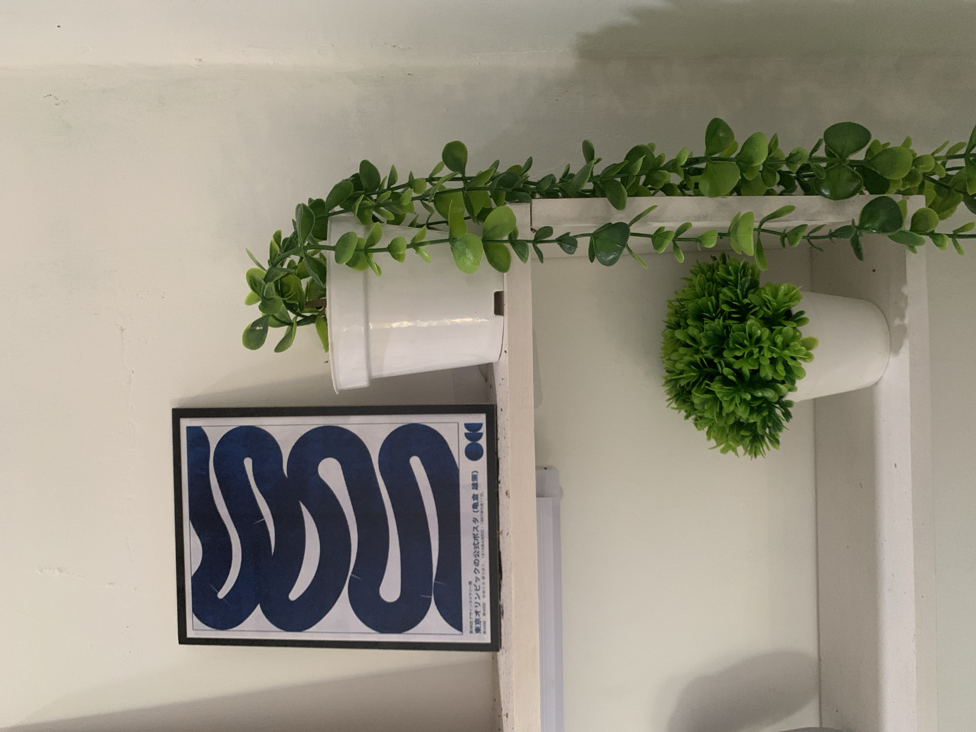 The image shows a shelf against a white wall. On the shelf, there is a framed poster with a blue abstract design and Japanese text. To the right of the poster, there is a white pot containing a trailing green plant. Below this, another white pot holds a small, bushy green plant. The lighting suggests an indoor setting, possibly a home or a decorative display. The location in Dukuh Mojo, Indonesia, is not depicted in the image and cannot be inferred from the visual content.