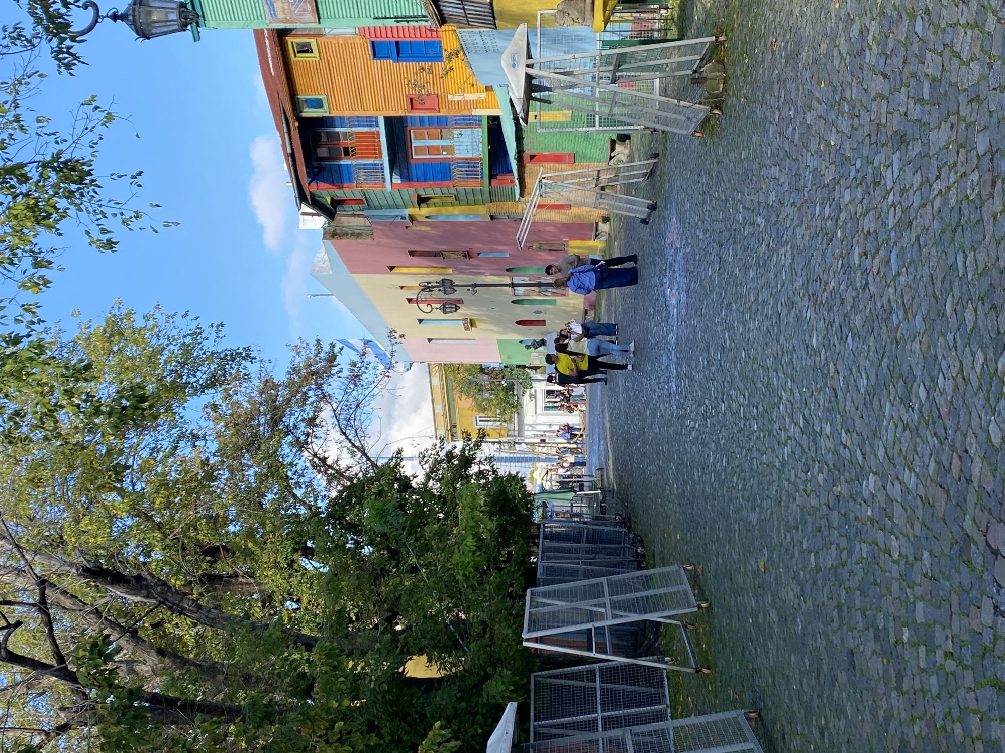 This vibrant image captures a picturesque street scene in El Caminito, La Boca, Buenos Aires, Argentina, characterized by its distinctive colorful architecture.
The scene is dominated by a row of multi-colored buildings lining the right side of a cobblestone street. These structures feature corrugated iron walls painted in bright hues of orange, blue, green, pink, and yellow, with contrasting window frames and balconies. On the left, lush green trees with dense foliage extend towards the sky, obscuring parts of the view. A classic-style black street lamp is visible in the upper left corner, extending from above the trees.
On the cobblestone street, several groups of people are seen walking and observing the surroundings. They appear to be tourists or visitors, casually dressed. Along both sides of the street, numerous metal barriers or fences, some appearing to be on wheels, are stacked or positioned. These may indicate crowd control measures, preparations for an event, or market stalls.
The setting is outdoors on a clear, sunny day, suggested by the bright blue sky with a few scattered white clouds. The natural lighting indicates it is likely mid-morning or early afternoon. The overall atmosphere is lively and cheerful, reflecting the iconic, artistic nature of the La Boca neighborhood. No discernible text is clearly visible within the image.