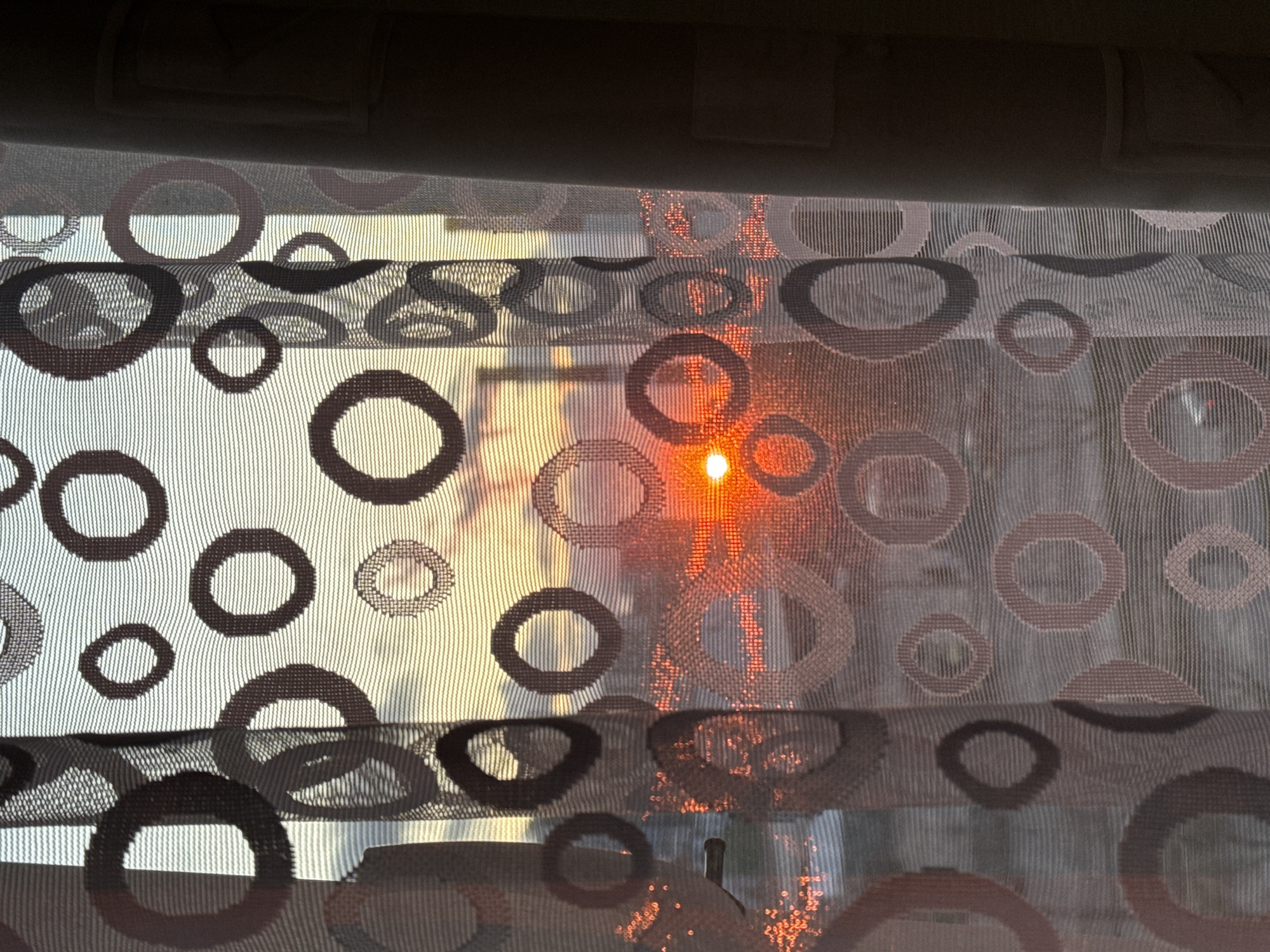 The image captures a view through a translucent window curtain adorned with a repetitive pattern of various-sized circles. The curtain itself appears to be made of a sheer, textured fabric with fine vertical striations. The circles are predominantly in shades of brown and dark grey, with some appearing as outlines and others as solid shapes.
A bright, intense point of light, presumably the sun, is visible towards the right side of the frame, piercing through the curtain and creating a vibrant orange and yellow glow. This suggests either sunrise or sunset. The light creates small, sparkling highlights on the curtain fabric, particularly in the area around the sun. Behind the curtain, the bright light illuminates the scene, giving hints of an outdoor environment, though no distinct details like buildings or landscape features are clearly discernible due to the curtain's opacity and the intense light.
At the top of the image, a darker, solid valance or curtain rod is visible, obscuring the uppermost part of the window. There are no people visible in the image, nor any discernible text. The scene conveys a sense of warmth and natural light entering a room in Saint Petersburg, Russia.