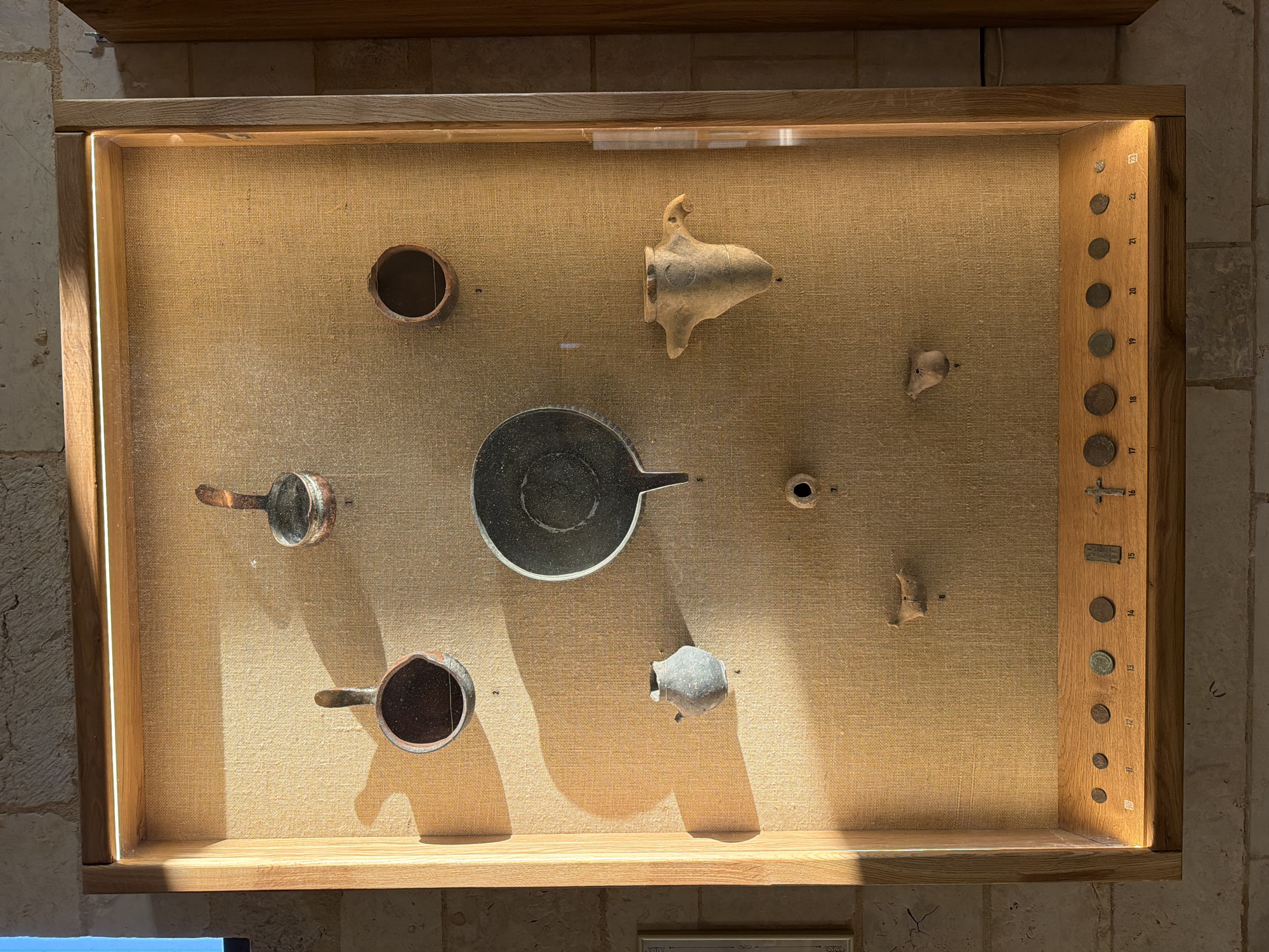 The image shows an exhibit in a museum display case. The display case has a light-colored wooden frame and a burlap-covered backing. Several ancient artifacts are mounted on the backing, including pottery shards, a small vase, and what appears to be a bronze ladle or scoop. There are also several coins and a cross displayed on a wooden shelf at the bottom of the case, each with a number below it, indicating an inventory or catalog system. The artifacts are illuminated by a light strip at the top of the display case, casting shadows on the burlap. The wall behind the display case is made of rough stone or brick. The image does not show the city of Скорняково, Russia.