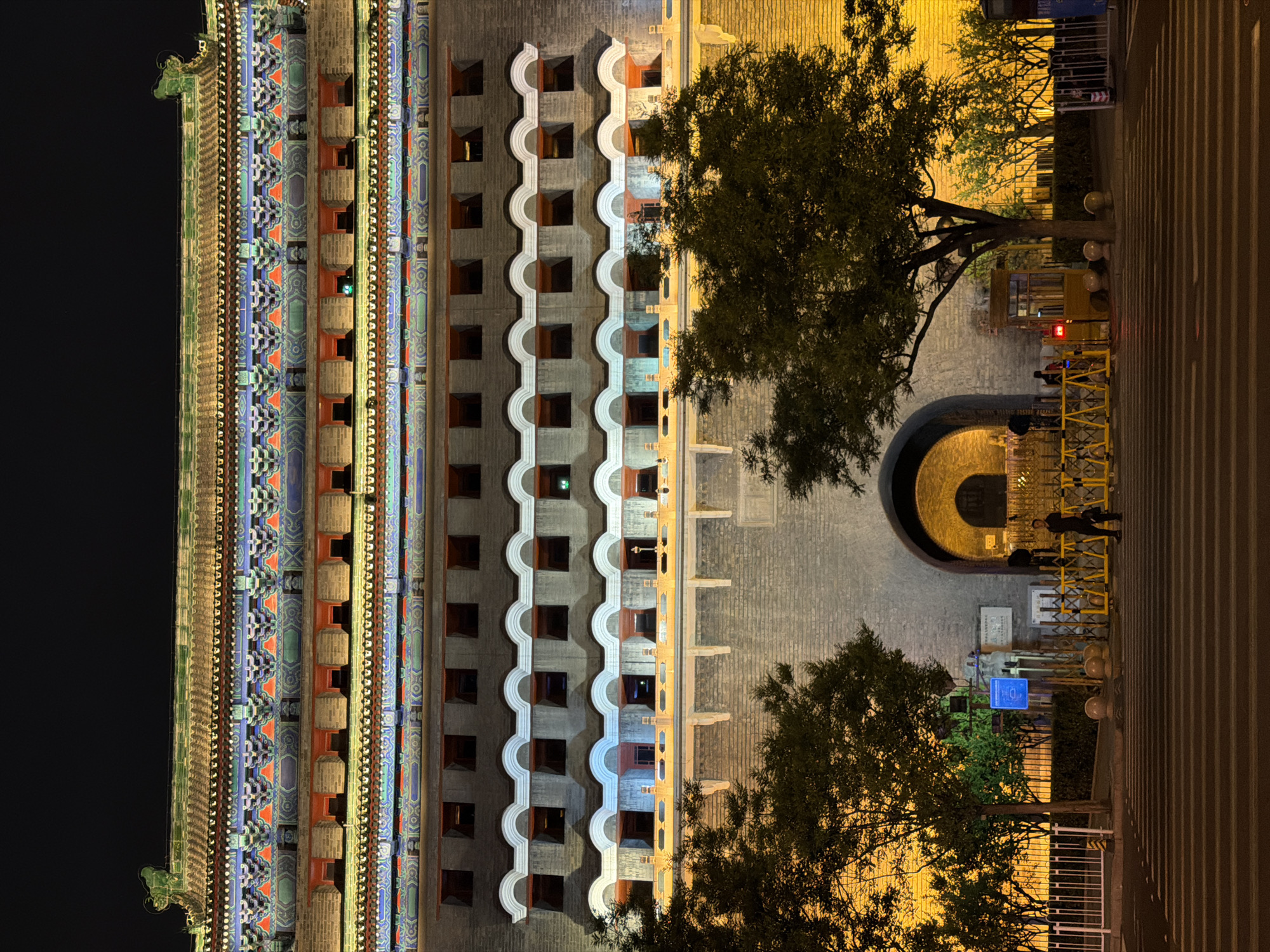Here is a detailed description of the image:
The image presents a night view of what appears to be the Gate of Supreme Harmony (Taihemen) in Beijing's Forbidden City, although this cannot be confirmed without additional information. The structure is seen from a high angle, potentially from an adjacent building. The left side of the image displays a part of the ornate, traditional Chinese architecture characterized by multiple tiers of intricately decorated eaves and roof structures, illuminated by artificial light that enhances its colors.
Adjacent to the building, a courtyard area is visible, partially lined with trees. A dark tunnel-like passage is visible, beyond which there are pedestrian barriers and what seems to be a security booth or checkpoint. Two figures appear to be present near this checkpoint. The courtyard is uniformly paved and well-lit.
The right side of the image shows a pedestrian area, with a set of steps leading away from the main structure and the courtyard. The scene is mostly illuminated, highlighting the details of the building and the courtyard, contrasting against the darkness of the night sky on the left. The overall setting is consistent with a major cultural or historical site that is open to the public.