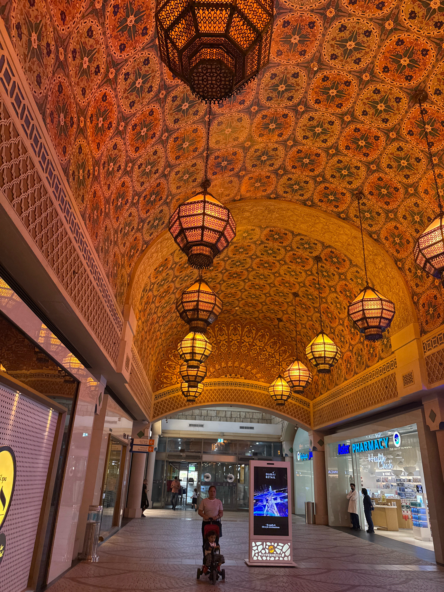 The image depicts a richly decorated interior space, likely a mall or a public building, with an ornate ceiling featuring intricate patterns and multiple hanging lanterns. The architecture suggests a Middle Eastern influence, possibly in Dubai, United Arab Emirates, due to the elaborate decorative style.
In the foreground, a woman is pushing a stroller with a child in it. They are walking down a tiled pathway. To the left, there are storefronts with glass facades and signage, one appearing to be for a spa. To the right, a brightly lit pharmacy is visible, with shelves stocked with products and two individuals, possibly employees or customers, standing near the entrance. A digital display screen is positioned in the walkway between the woman with the stroller and the pharmacy, showing an advertisement. The overall lighting is warm, emanating from the lanterns and interior lights of the shops. The ceiling is the dominant decorative element, with repeating floral or geometric motifs in shades of orange, yellow, and blue.