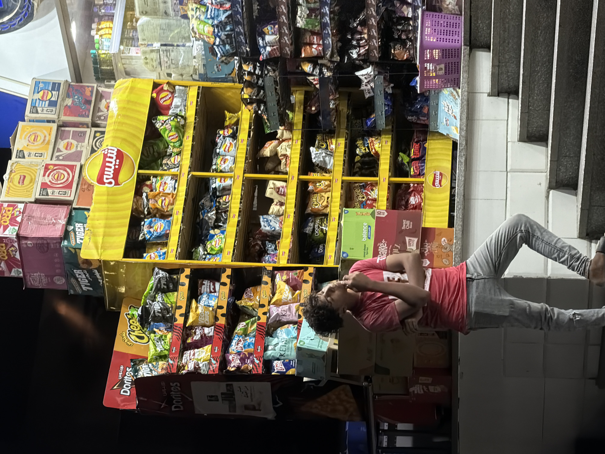 The image depicts a young person standing on a set of stairs in front of a brightly lit store, likely a convenience store or small supermarket. The store appears to be well-stocked with various snack items, predominantly chips and packaged goods, displayed on yellow and dark shelves. The shelves are filled with an assortment of colorful bags and boxes, featuring brands such as Doritos and Cheetos, as well as other unidentifiable snacks. The lighting suggests it is either nighttime or an indoor setting with artificial illumination. The person is wearing a red t-shirt and gray jeans, with their arms crossed and looking upwards. The background shows more shelves of products and a glimpse of what might be a street or exterior beyond the store. The overall scene gives the impression of a casual moment captured outside a local shop in the city of 6th of October, Egypt.