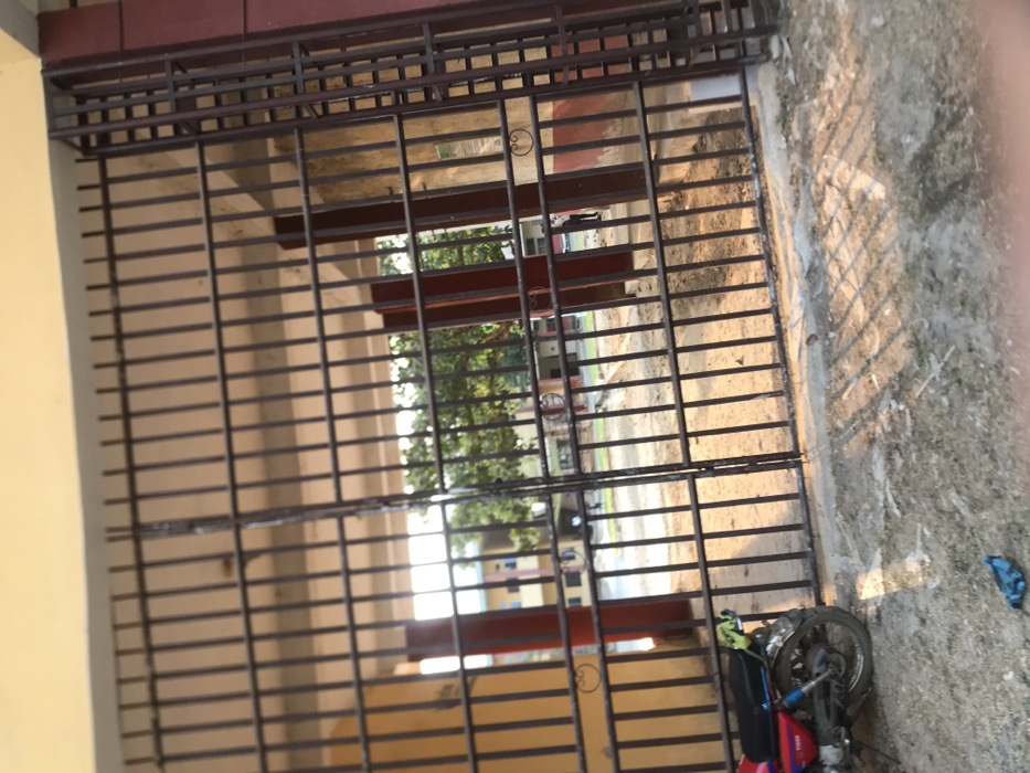 This image captures a ground-level view through a large, dark metal gate, likely an entrance or perimeter barrier, in Umudike, Nigeria. The gate features a grid pattern of vertical and horizontal bars. To the immediate left, a light yellow-colored wall abuts the gate structure, with a darker, reddish-brown band above it.
The foreground ground directly outside the gate, on the right side of the image, is unpaved, appearing dusty, sandy, and somewhat uneven. Strong, elongated shadows from the gate bars are cast across this ground, suggesting direct sunlight and either early morning or late afternoon. A red and black motorcycle is lying on its side on this dusty ground in the bottom right corner, with its front wheel and parts of the engine visible. A small blue object, possibly cloth or plastic, rests on the ground further to the right.
Beyond the gate, green trees with dense foliage are visible, indicating an outdoor, possibly green space. Further in the background, past the trees, light-colored buildings with some darker accents can be seen, hinting at a distant residential or institutional complex. The bright light passing through the gate suggests clear, sunny weather. No people or discernible text are visible in the scene, and no specific activity or event is taking place, presenting a still, observational view.