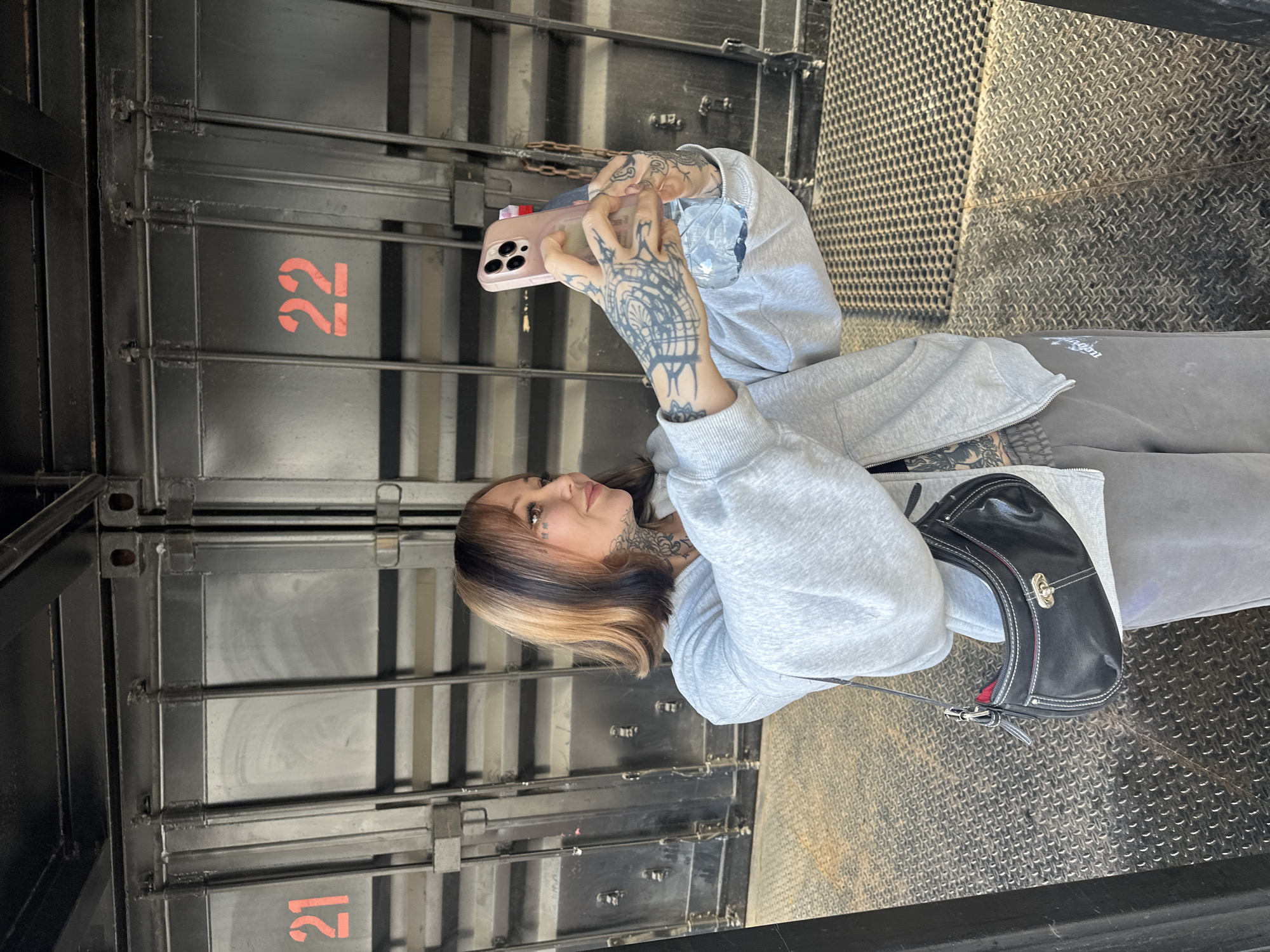 A woman with blonde and dark hair, wearing a gray hoodie and gray sweatpants, is standing in what appears to be a metal-walled structure, possibly a freight elevator or loading bay. She's holding a pink smartphone and taking a picture or video, with a partially visible plastic water bottle nearby. The woman has extensive tattoos on her hands, arms, and neck. A small black purse hangs from her shoulder. The walls of the structure are metal with a grid pattern on one wall and numbered panels on another. The numbers '21' and '22' are visible on these panels. Based on the architectural style and the numbered panels, this location is very likely to be a loading bay or freight elevator within an industrial area of London, United Kingdom.