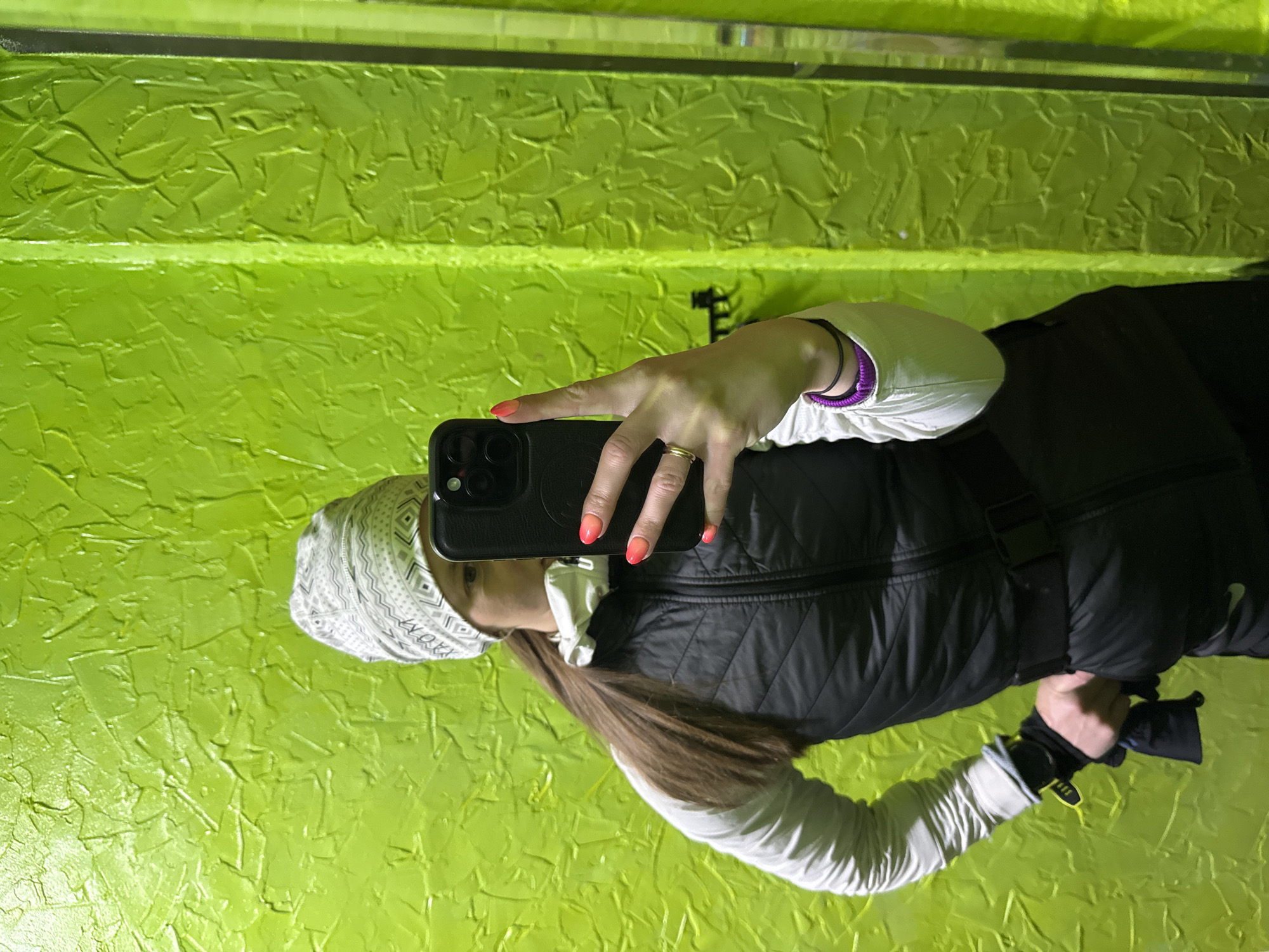 A woman in winter attire takes a selfie indoors. She wears a white and gray patterned beanie, a black puffer vest, and a white long-sleeved shirt with a purple-trimmed cuff. Her nails are painted with a pink ombre effect. She holds a black smartphone in her left hand. She wears a gold ring on her ring finger. Part of her brown hair is visible. The background is a bright green textured wall. The location is unknown, and the photo provides no direct evidence to suggest it was taken in the city of Chaikovsky, Russia.