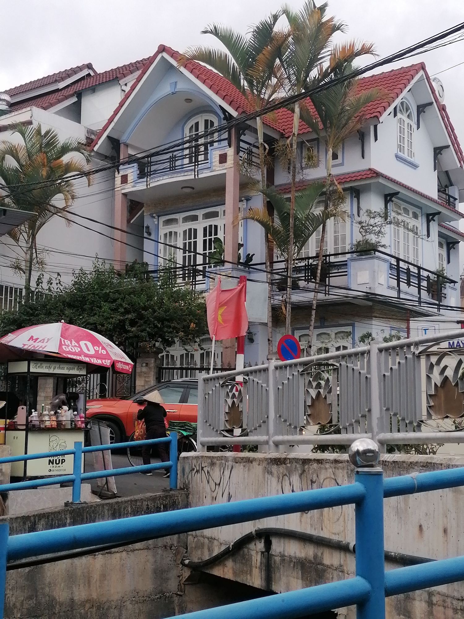 The image captures a street scene in Cam Ly - Đà Lạt, Vietnam. In the background, there is a multi-story building with red tile roofs and a blue and white facade. Palm trees stand tall in front of the building, partially obscuring the view of the architecture. Power lines crisscross the sky above.
In the midground, an orange car is parked near a street-side stall or cart with a pink and white umbrella. The umbrella has text in Vietnamese: "MAY TỈNH," "GOP 0%," "NGUYỄN VĂN CỪ chore," and "Hello, café gi chuta nguoi dep." Next to the stall, a person wearing a conical hat is bent over a bicycle. A red flag is visible behind the stall. A blue and red 'no parking' sign is also present.
In the foreground, a blue metal railing separates the street from what appears to be a lower level, possibly a waterway or a sidewalk. The barrier and the concrete surfaces below show signs of wear and graffiti. A sign with "NÚP" and a phone number is visible.