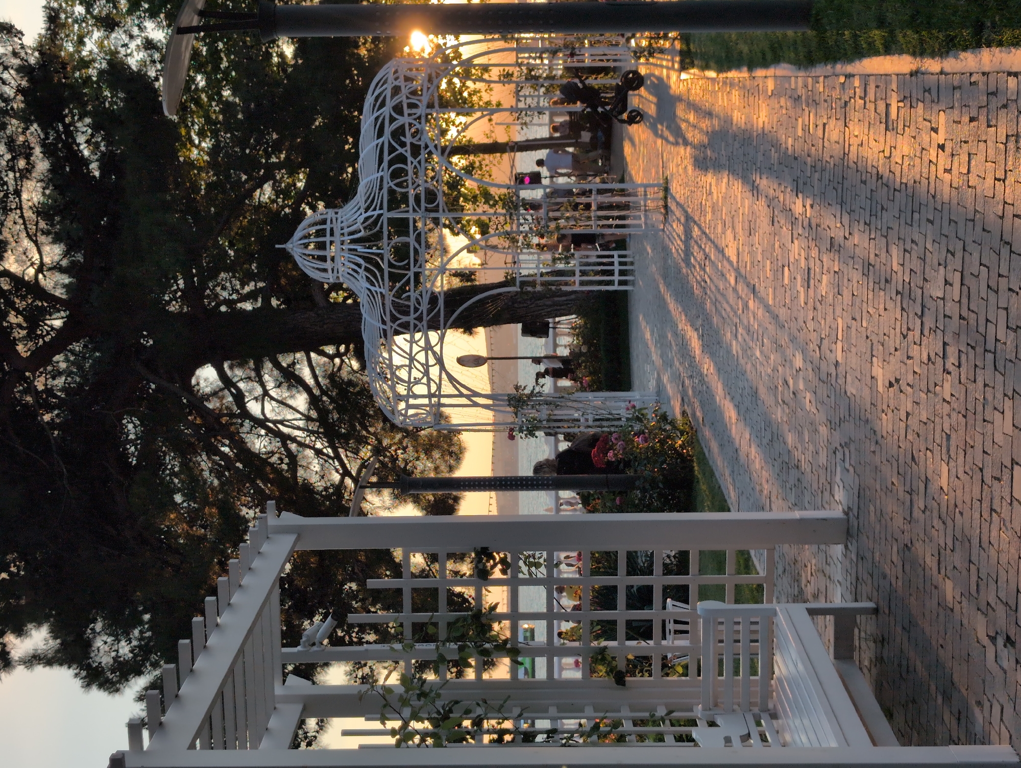 The image depicts an outdoor scene at sunset, likely in a park or promenade near a body of water. In the foreground, a white wooden pergola with intricate latticework extends into the scene, partially obscuring a white bench. The ground is paved with reddish-brown bricks, and long shadows are cast across the path by the setting sun, which is visible through the trees and structures in the background.
Several people are visible in the middle ground, some standing near the water's edge and others near a white, ornate gazebo. A baby stroller is parked near the gazebo. Lush green foliage and flowering bushes are present along the path and around the structures. In the distance, a body of water is visible, with a hint of a shoreline or distant structures. The overall atmosphere is serene and bathed in the warm, golden light of sunset. The location is unknown.
