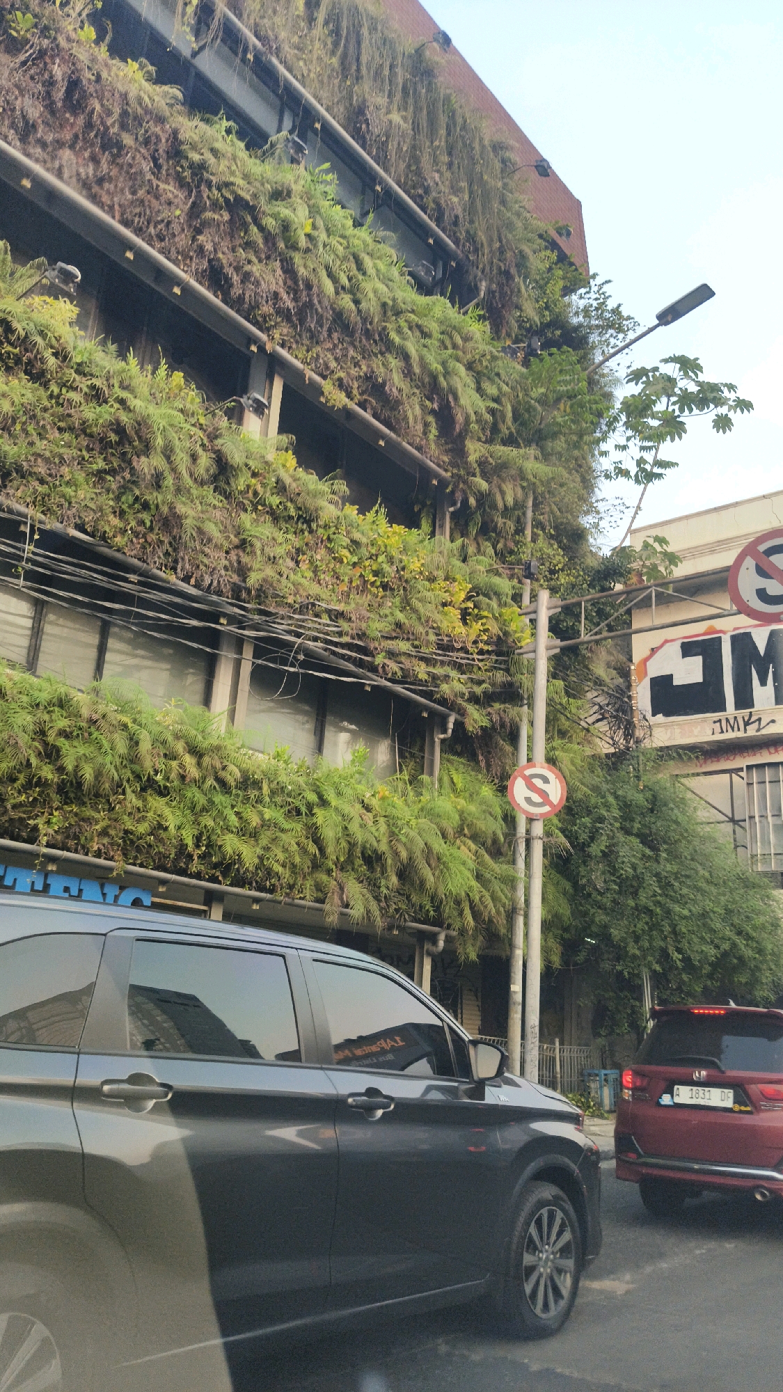 A medium shot captures a building facade covered in lush greenery, suggesting a vertical garden. The building has multiple stories with windows and some visible infrastructure like electrical wires and lighting fixtures. On the right side of the image, a red car with a license plate reading "A 1831 DF" is visible, stopped behind a dark gray SUV in the foreground. A streetlamp and traffic signs, including a circular sign with a red border and a diagonal line, are also present. The overall scene appears to be a street in a city, possibly Central Jakarta, Indonesia, given the context.
