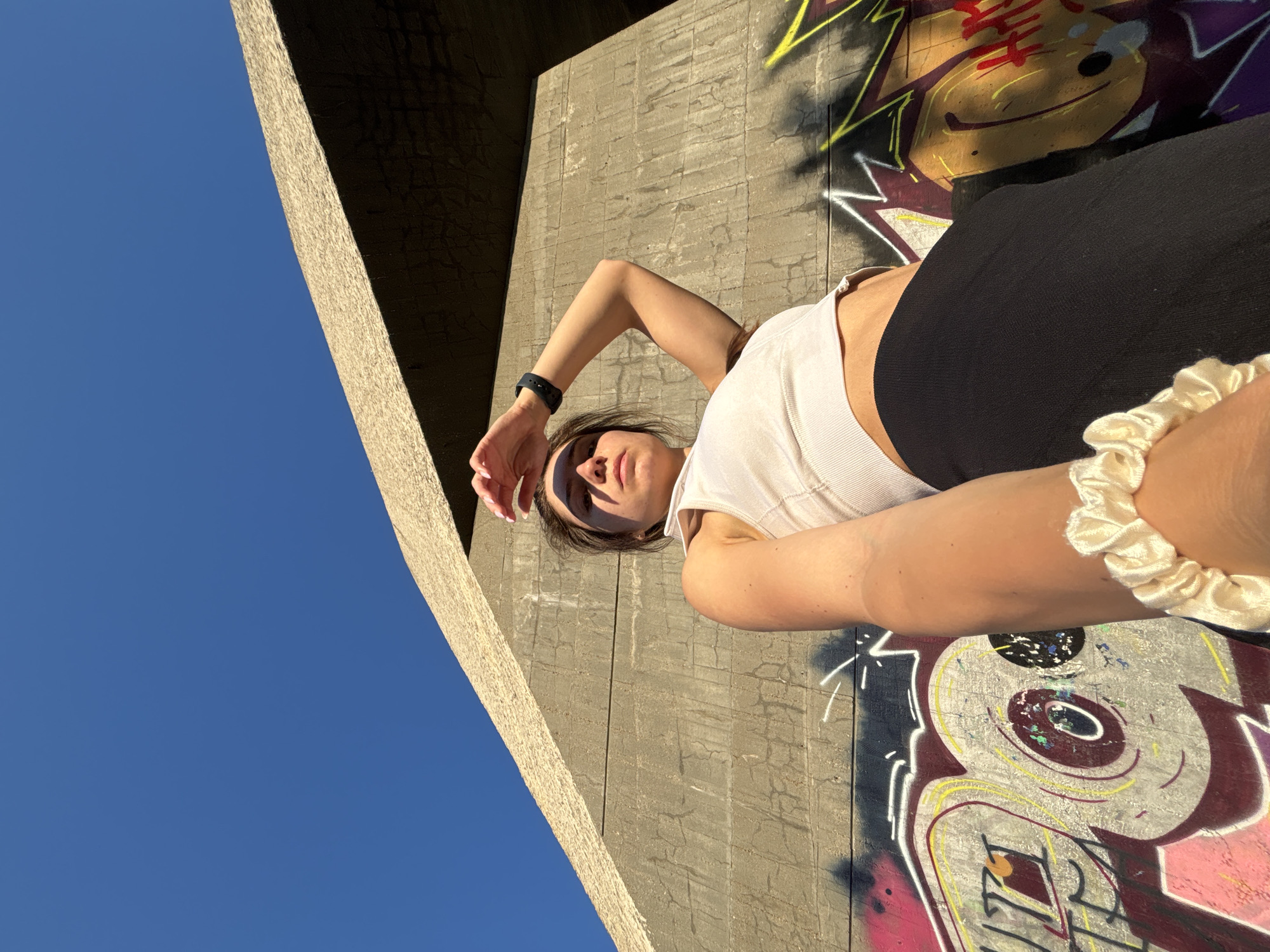 A low-angle, outdoor, eye-level, medium shot captures a young adult woman standing outdoors in Varna, Bulgaria. The woman, facing the camera, has her right arm raised to shield her eyes from the sun, and her left arm extends towards the camera, holding it. She is wearing a white cropped tank top and a black skirt. Behind her, a rough, concrete wall is covered in vibrant graffiti. The wall meets a textured, sloping concrete structure that angles upward toward a clear, bright blue sky. The lighting suggests it is daytime, with the sun casting sharp shadows.