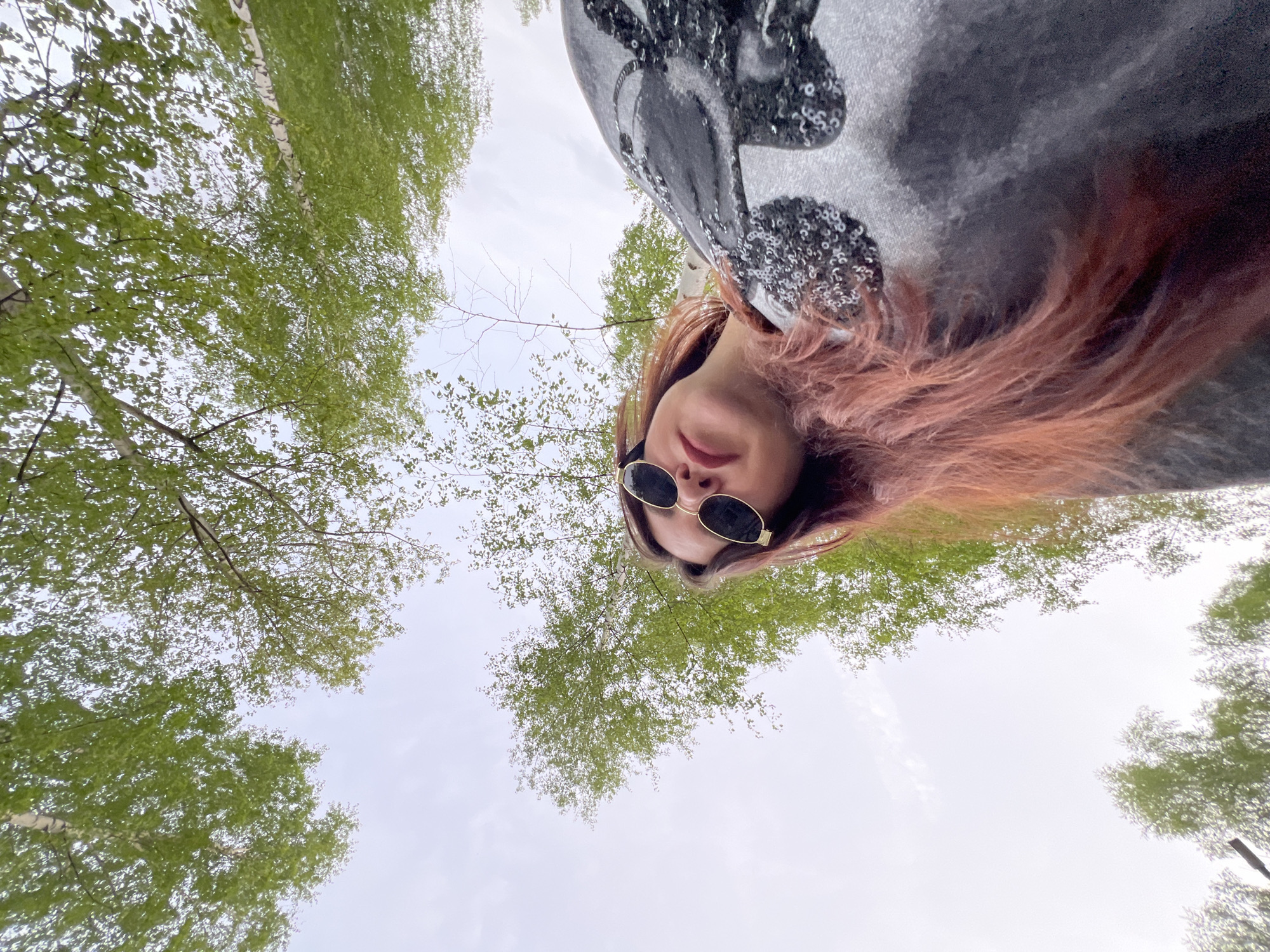 A low-angle, upward-looking photograph shows a person wearing sunglasses and a gray top with sequined Mickey Mouse ears, positioned against a backdrop of trees with green leaves. The person's reddish-brown hair is visible around their face. The trees appear to be birch, suggested by the white bark, and fill the perimeter of the frame against a light sky. The location is potentially near birch trees in the city of Noviy, Russia, if that information is relevant to the image's context.