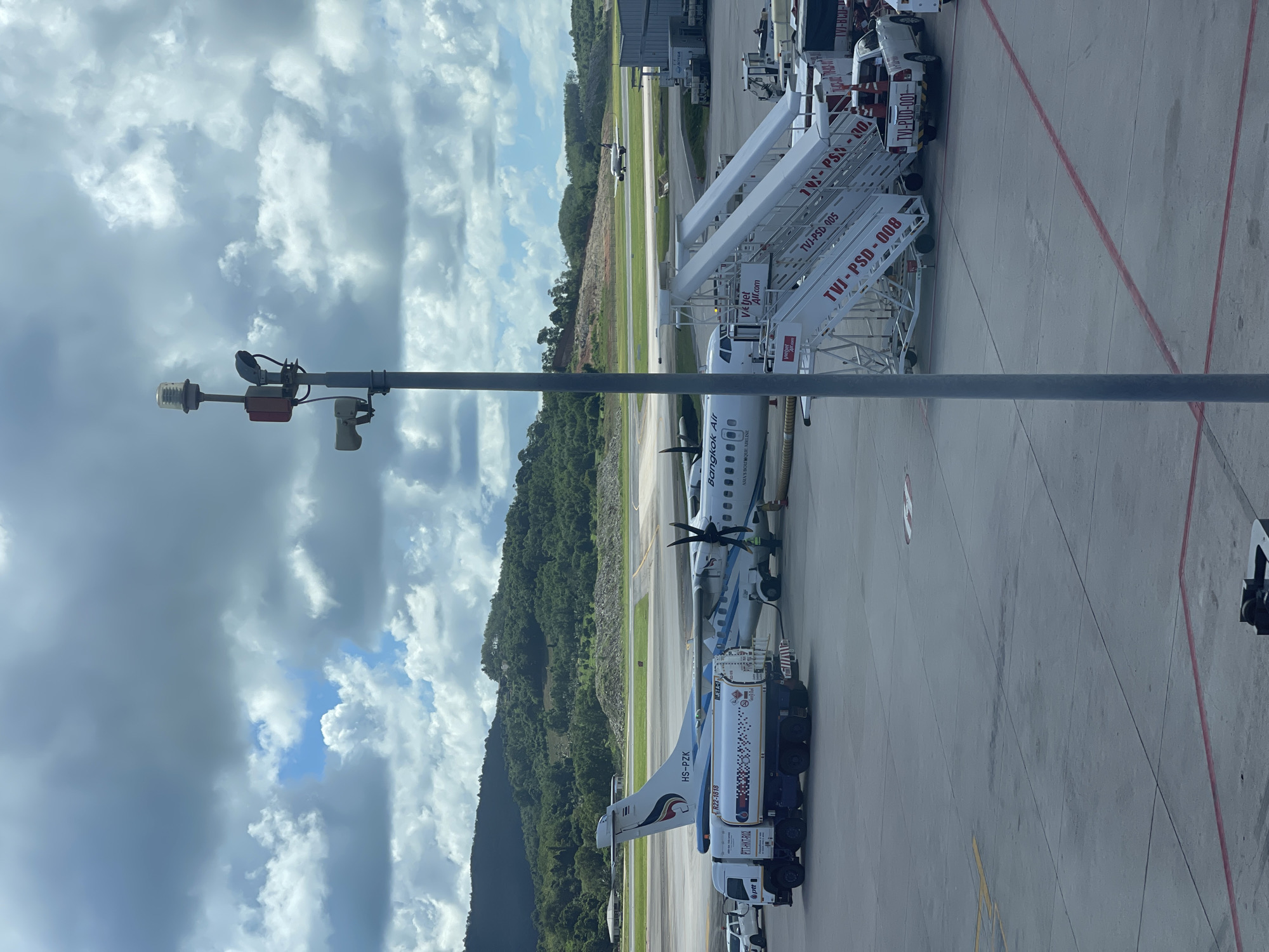 This image captures an airport scene in Mai Khao, Thailand, under a partly cloudy sky. A Bangkok Airways turboprop aircraft, identified by the registration "HS-PZK" and the airline's logo, is parked on the tarmac. A fuel truck, labeled "PTT-HKT-R02," is connected to the aircraft, indicating refueling operations are in progress. Mobile stairs, marked with "Vietjet Air" and various codes such as "TVI-PSD-005" and "TVI-PSD-008," are positioned next to the aircraft. A white pickup truck is parked near the stairs. In the background, lush green hills are visible, and another aircraft is seen on a distant runway. A tall pole with lights and a camera is prominently featured in the foreground, obscuring part of the sky and landscape. The overall environment is that of an active airport apron.