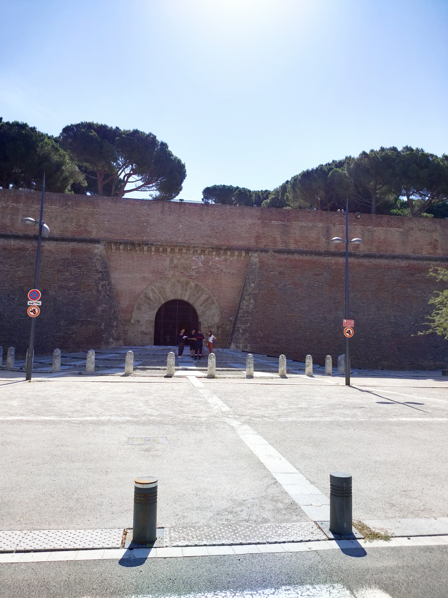 The image shows the entrance to Fort de Bug in Perpignan, France. The fort is characterized by a large brick wall with a semicircular arched entrance. Several people are gathered at the entrance, appearing to be engaged in conversation. Two lampposts stand on either side of the entrance, and traffic signs are visible near the left lamppost. The ground in front of the fort is paved, with bollards lining the area. Trees with dark green foliage are present behind the wall, and the sky is clear and blue.