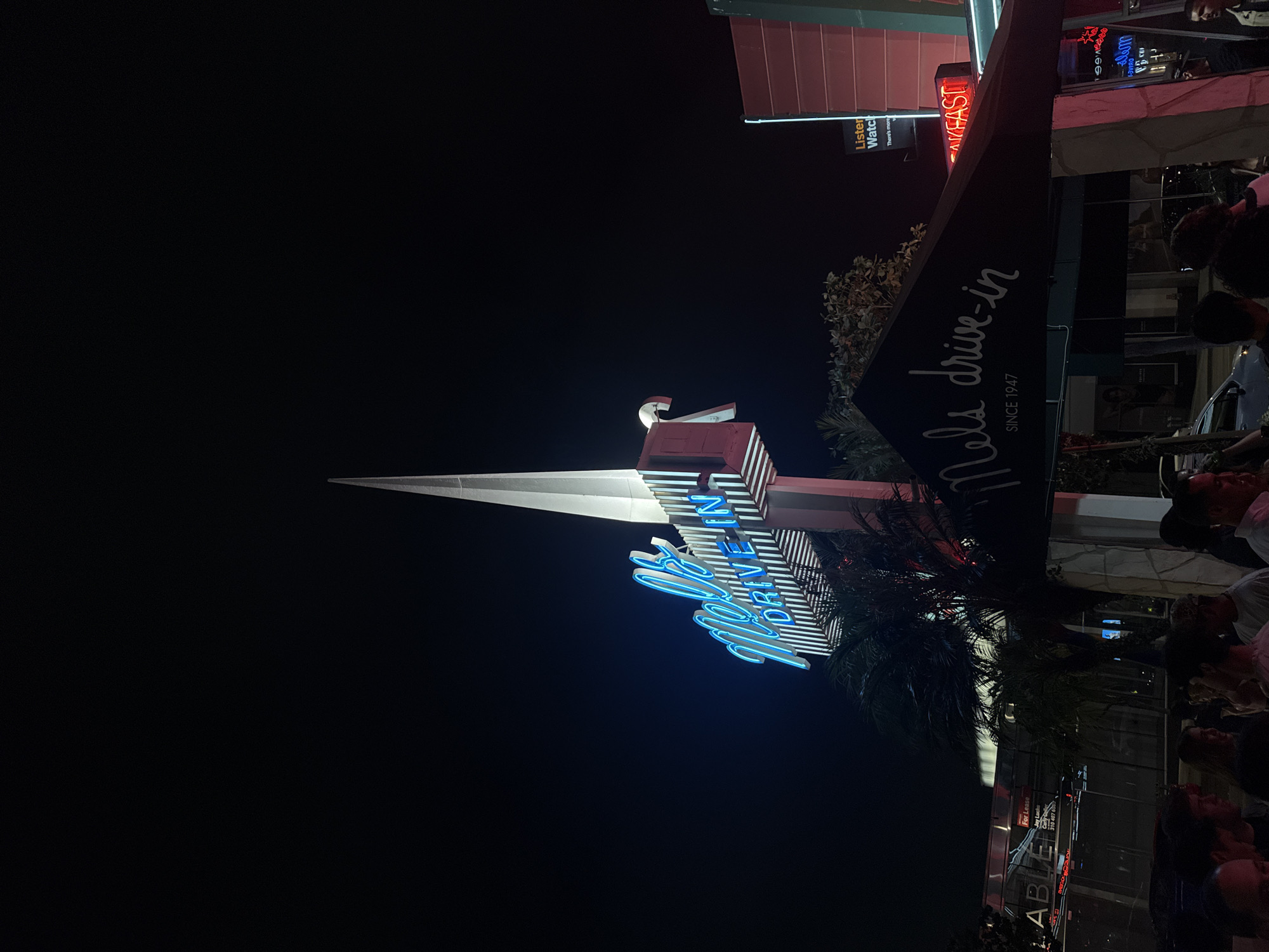 The image showcases the "Mel's Drive-In" neon sign, taken at night in West Hollywood, USA. The sign is illuminated in blue against the dark sky. A large black sign displaying "Nels drive-in SINCE 1947" is visible. Nearby signs read "BREAKFAST", "Listener Watch", and "A/FAST". A building with a 'For Lease' sign reading 'ABLE' and a phone number is also visible. People are situated around the area. Palm trees provide an ambient element to the environment.