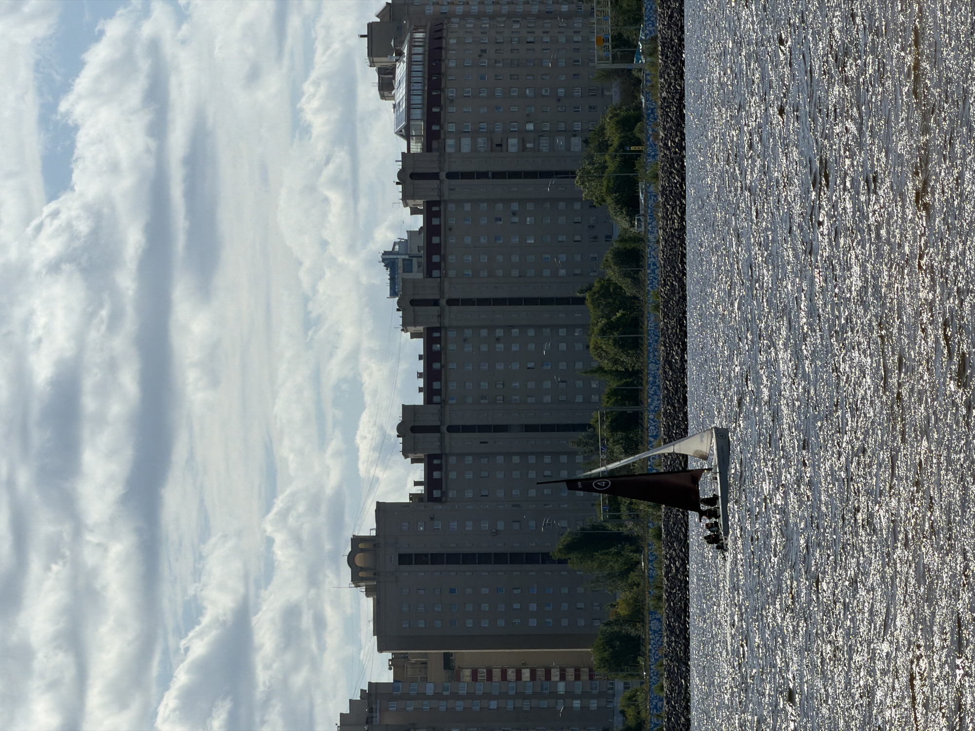 A sailboat with a dark red sail and a white jib sails on the water. The sailboat has several figures on board. In the background, a row of large, multi-story apartment buildings lines the shore, with trees and a road visible between the water and the buildings. The sky is filled with dramatic clouds. This scene is consistent with the appearance of the area along the Gulf of Finland in Saint Petersburg, Russia, specifically near the residential developments along the Primorsky District's coast.
