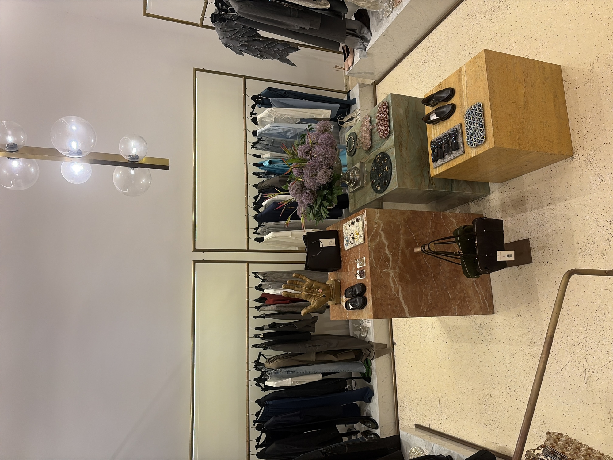 The image shows the interior of a modern boutique in Tbilisi, Georgia. The boutique features a clean, minimalist aesthetic with light-colored walls and floors. Clothing racks are organized with various garments, primarily in neutral tones like black, gray, and white. Several display stands are strategically placed throughout the space, showcasing accessories.
On the left side of the image, a display stand made of red marble holds a golden hand sculpture with a necklace, a black handbag, and a pair of black loafers. Next to it, a smaller green marble stand displays a floral arrangement and several clutches or small bags.
On the right side, a light wood cube display stand holds a pair of black loafers, sunglasses, and a patterned accessory. In the background, clothing is displayed on racks, with one rack featuring a distinctive grey garment with layered, wing-like elements. A modern chandelier with multiple glass globes hangs from the ceiling. The overall impression is of a high-end fashion boutique.