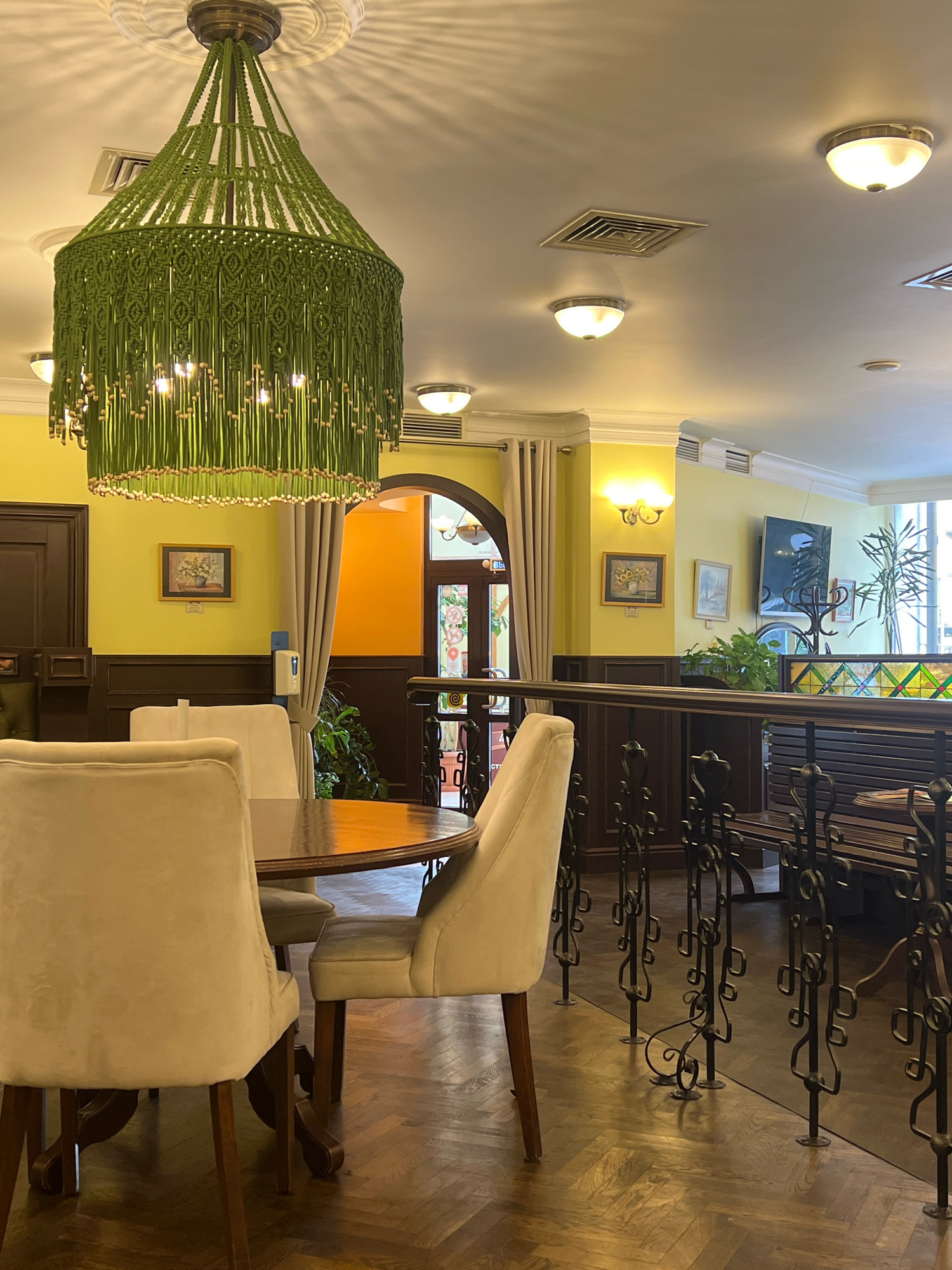 The image shows a well-lit interior of a restaurant, possibly located in Kaliningrad, Russia, given the Cyrillic text visible on a door in the background. The foreground features a round wooden dining table with three light-colored upholstered chairs around it. The table appears to be polished and reflects the light. A decorative, green macrame chandelier hangs from the ceiling, illuminating the scene. The walls are painted a pale yellow and are adorned with framed pictures. A dark wooden banister with ornate metalwork separates the dining area from another section of the restaurant, which includes a bench seat and decorative stained glass. Several ceiling lights and air vents are visible on the light-colored ceiling. The floor is made of dark wooden planks laid in a herringbone pattern. The overall impression is one of a cozy and somewhat upscale dining establishment.