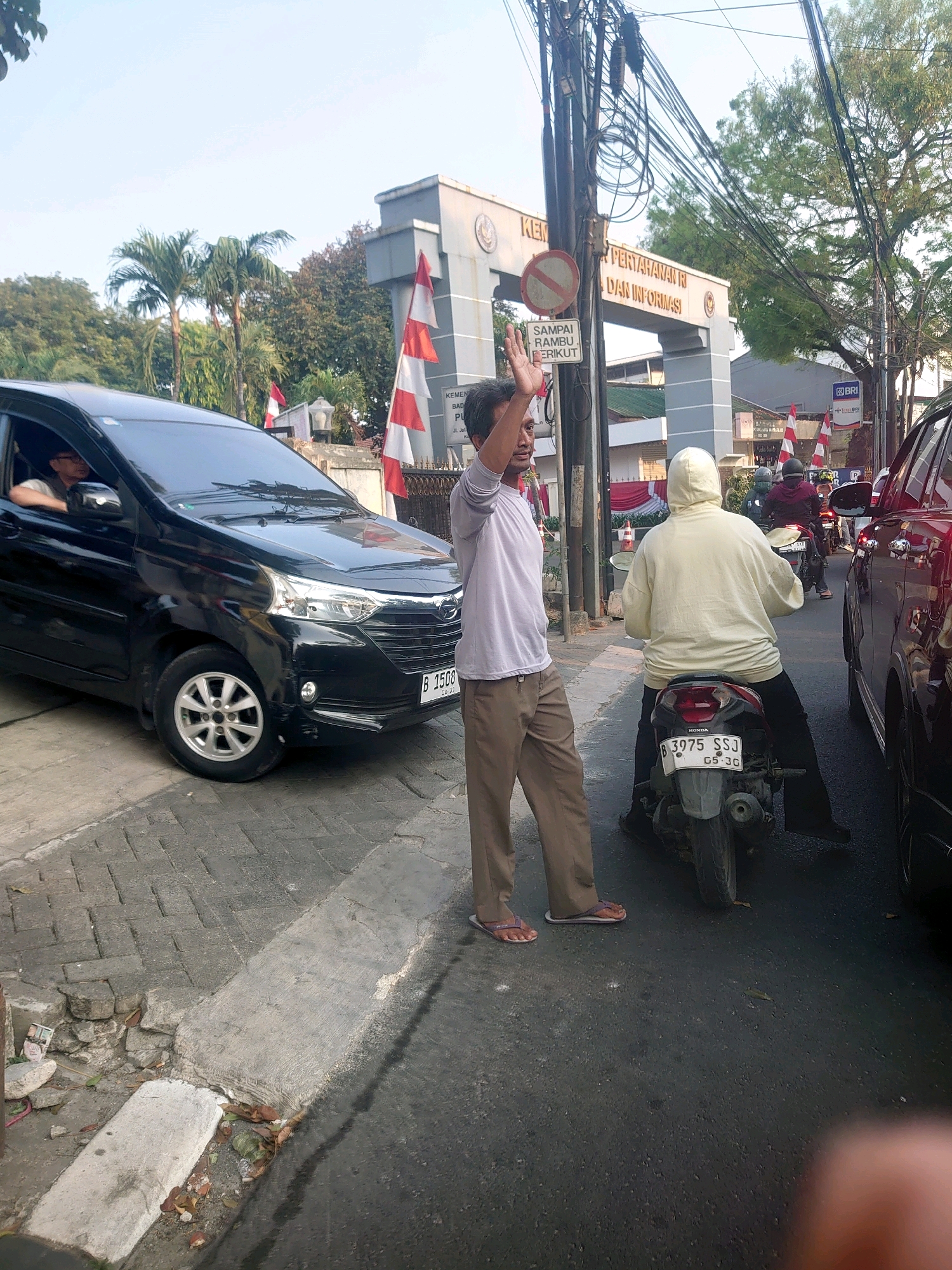 The image depicts a street scene in South Jakarta, Indonesia, near an entranceway to a government building labeled "KEMENTERIAN PERTAHANAN RI DAN INFORMASI" (Ministry of Defense and Information). A black car is parked on the left, with a driver visible inside. A man in a white shirt and brown pants stands on the sidewalk, waving. In the street, a person in a light-colored hooded jacket is riding a motorcycle. Another motorcycle and a red car are visible further down the street, suggesting traffic. Indonesian flags are displayed prominently, both on poles and attached to the building. The environment appears to be urban, with paved roads, sidewalks, streetlights, and trees. The overall impression is of a busy street with government activity in the background.