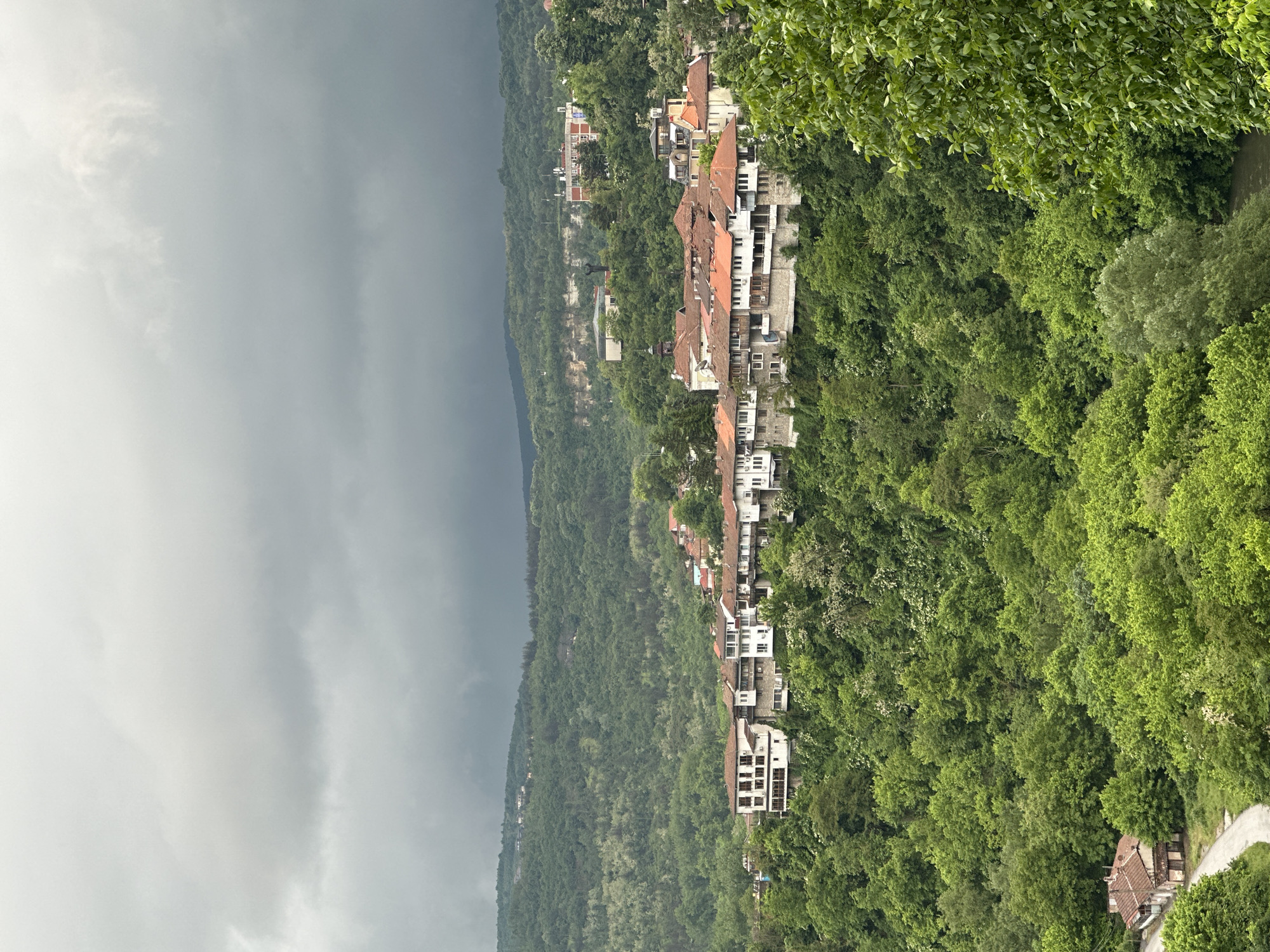 The image shows a view of Veliko Tarnovo, Bulgaria, likely taken from a high vantage point. The scene depicts a steep, forested hillside with residential buildings constructed along its slope. The buildings are a mix of white and light-colored structures, some with red-tiled roofs. The hillside is covered in dense green trees. In the background, a dark, ominous sky suggests an approaching storm. A road winds along the lower edge of the frame.