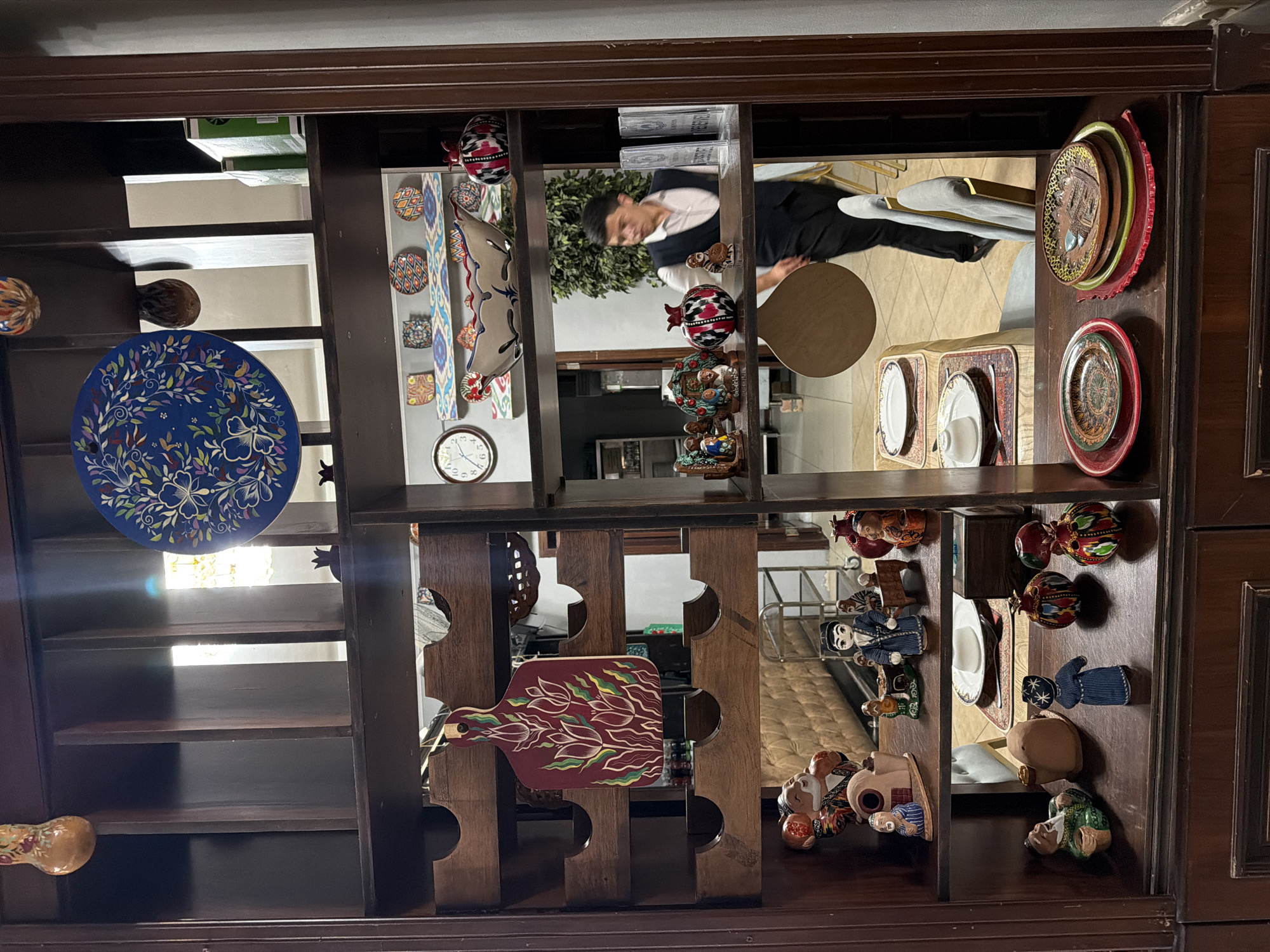 The image captures a dimly lit, traditional Uzbek restaurant interior, likely in Chinarkent, Uzbekistan, as suggested by the context. The foreground is dominated by a dark wooden shelving unit filled with decorative items. On the left side of the upper shelf, a wooden cutting board with a red base and white and green floral motifs is displayed. The middle shelves are populated with various ceramic figurines and decorative pomegranates, some with intricate geometric patterns in red, white, and black.
On the right side of the middle shelves, a clock with Roman numerals is visible, along with decorative plates and a large, ornate ceramic bowl. A waiter, dressed in a white shirt and black vest, is partially visible on the right, walking behind the shelving unit, indicating an active restaurant environment. He is carrying a circular wooden object.
The lower shelves showcase more ceramic figurines, including one resembling a man in a green robe and another in a blue traditional outfit. Plates are stacked on the bottom right shelf, featuring colorful, intricate designs. The overall atmosphere suggests a cultural dining experience, with an emphasis on traditional Uzbek artistry and decor.