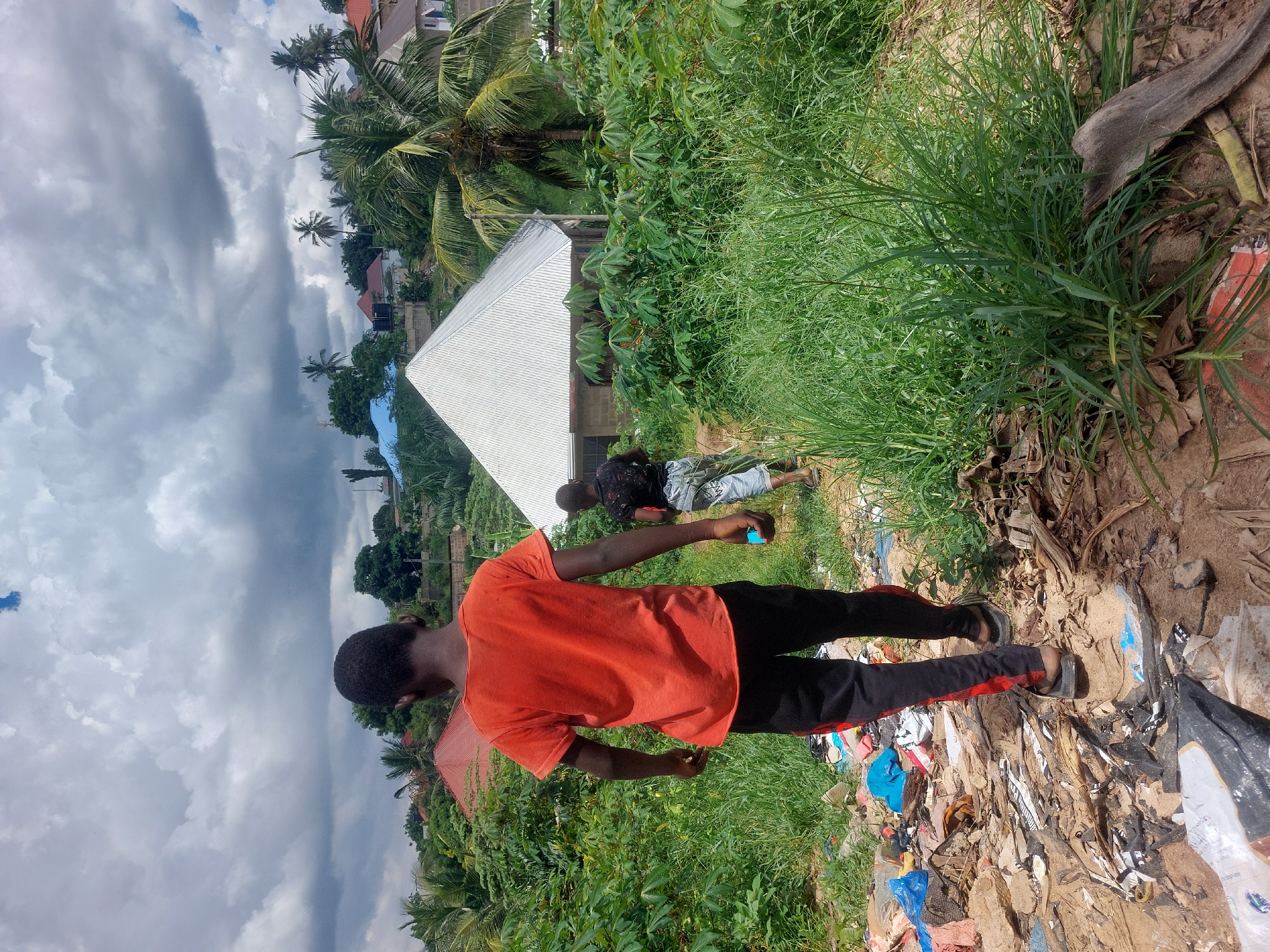 The image shows two young males outside in what appears to be a semi-urban or peri-urban environment. The foreground features a younger male wearing an orange t-shirt and black pants with a red stripe. He is standing on a patch of ground covered with trash, including plastic bags and debris. In the middle ground, another male is visible, wearing a patterned shirt and shorts. He appears to be handling a small blue object. Behind them, there is a building with a silver metal roof and green vegetation. In the background, there are more buildings, palm trees, and a cloudy sky. The scene suggests an area where residential zones interface with less formal waste disposal practices. Without more data or more objects, determining the exact location within Dar es-Salaam, Tanzania is not possible.