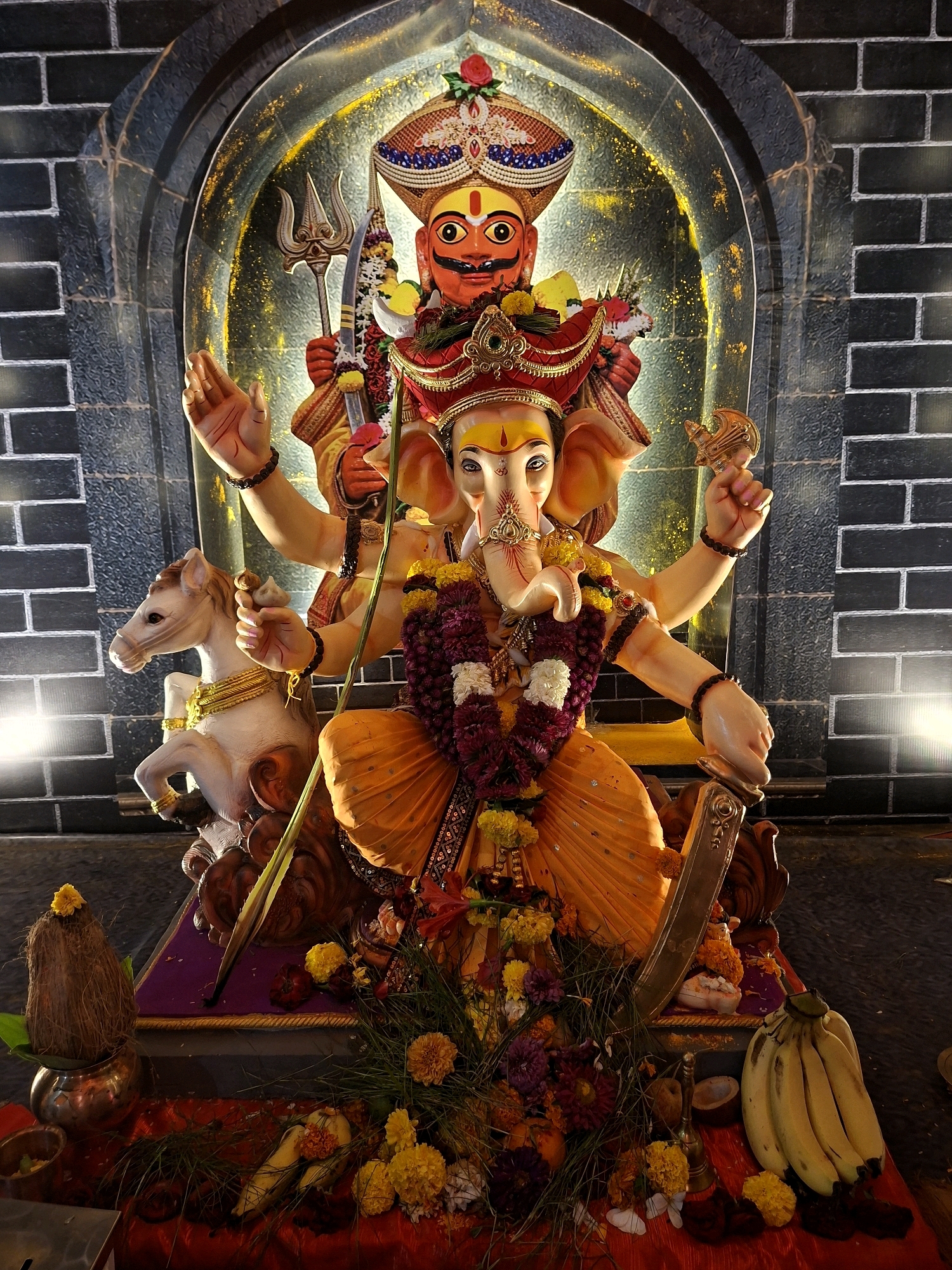 The image shows a devotional display of the Hindu deity Ganesha, accompanied by another figure, within an arched alcove. The figures are adorned with garlands of flowers, beads, and elaborate headdresses. Ganesha, a prominent deity with an elephant head, is depicted in a seated position, with multiple arms holding various objects, including a sword, a lasso, and a modak (a sweet dumpling). The accompanying figure, which appears to be a representation of a god with a human-like face and orange skin, is positioned behind Ganesha, holding a trident and a sword.
The figures are placed on a tiered platform decorated with a variety of flowers, including marigolds and roses, as well as bananas and a coconut. To the left of the platform, a white horse statue is partially visible. The background consists of dark brick walls and an illuminated arched opening with a golden, speckled pattern. Lighting emanates from the sides, casting highlights on the figures and the surrounding decorations.
The scene is indicative of a religious festival or shrine, possibly related to Ganesh Chaturthi, a popular Hindu festival celebrated in India. While the image itself does not explicitly state its location, similar elaborate displays are commonly found in temples and public spaces across cities like Pune, India, during such festivities. The craftsmanship of the idols, the floral decorations, and the overall presentation are characteristic of religious art and devotion prevalent in that region.