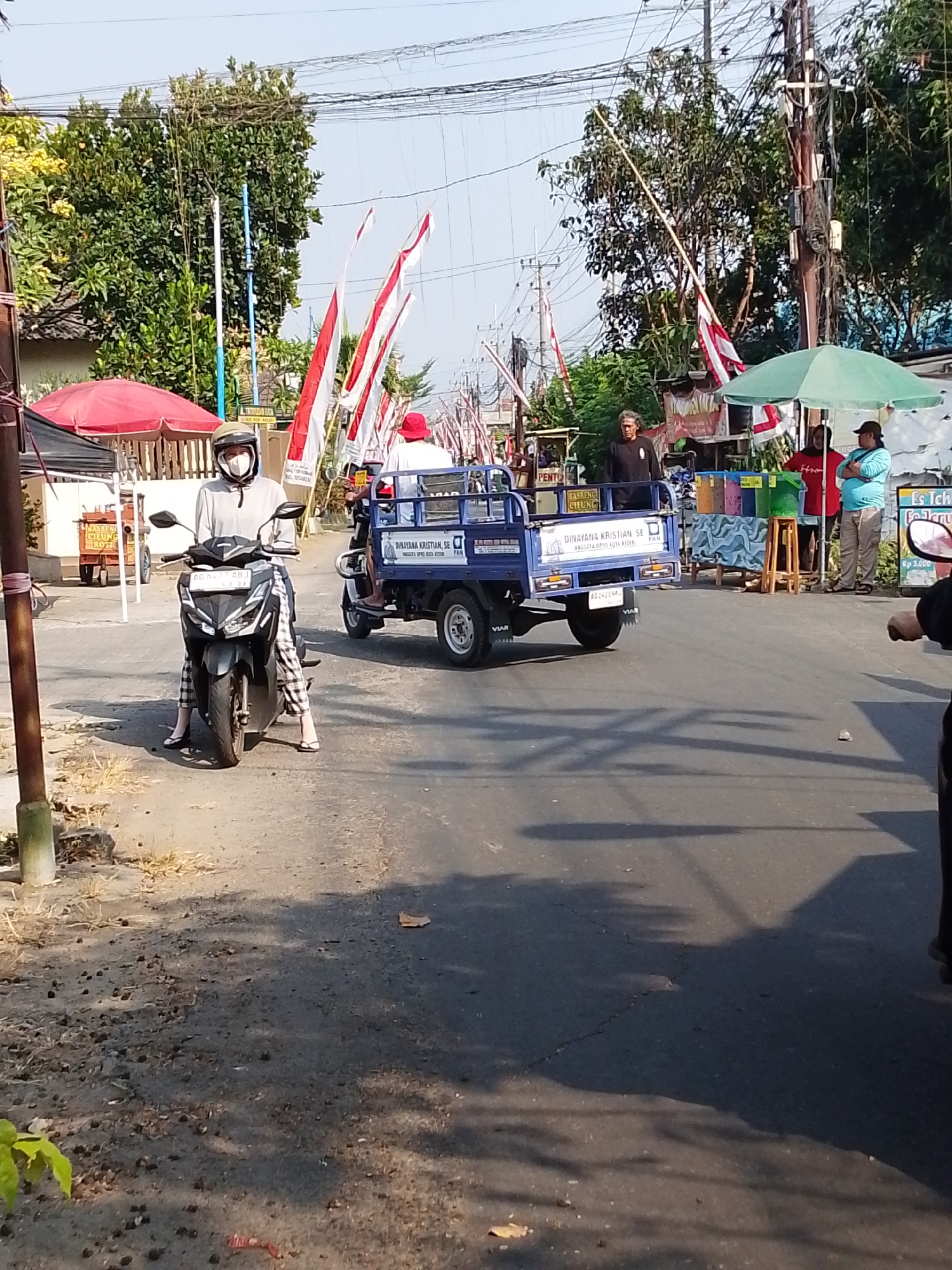 The image depicts a street scene in the city of Kediri, Indonesia, on a sunny day. In the foreground, a person wearing a grey hooded jacket, a helmet, and a face mask is seated on a black and silver motorcycle. The motorcycle has a license plate that reads "AG 6047 ABJ". The rider is facing forward, and their legs are crossed. The person is wearing checkered pants and white sandals.
Further down the street, a small blue truck with a canopy is moving. The truck has a sign on its side that says "DINAYANA KRISTIAN SE" and "ANGGOTA DPRD KOTA KEDIRI". There are two people in the truck bed, one wearing a white shirt and a red hat, and another person in the driver's seat wearing dark clothing.
On the right side of the street, there are several stalls or vendors. One stall has a green umbrella and is selling colorful items in buckets. Three people are visible near this stall: one wearing a dark shirt, another in a red shirt, and a third in a light blue shirt and a dark cap.
In the background, trees and utility poles with numerous wires are visible. Indonesian flags, consisting of red and white stripes, are prominently displayed on poles along the street, suggesting a national celebration or event. The road surface appears to be asphalt, with some patches of dry grass and dirt along the sides. The overall environment suggests a typical street in a residential or commercial area of Kediri City.