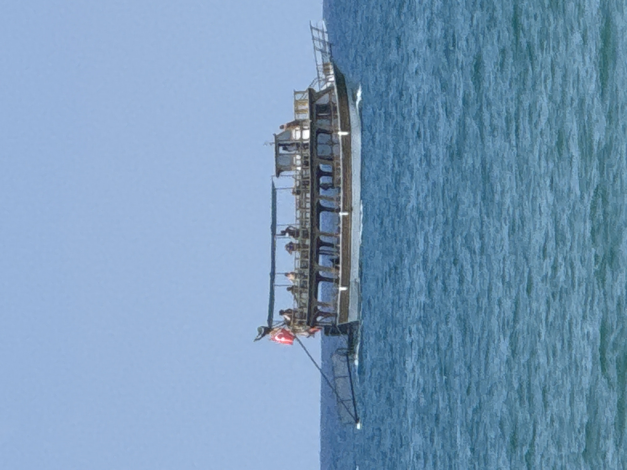 A wooden tour boat with a white hull and a brown upper deck is sailing on the blue sea under a clear sky. The boat is carrying several passengers on its open upper deck, some of whom are seated under a canopy. A Turkish flag is visible on the stern of the boat. The boat appears to be in motion, as indicated by the wake behind it. The sea is calm with small waves. This image was taken in Manavgat, Turkey.