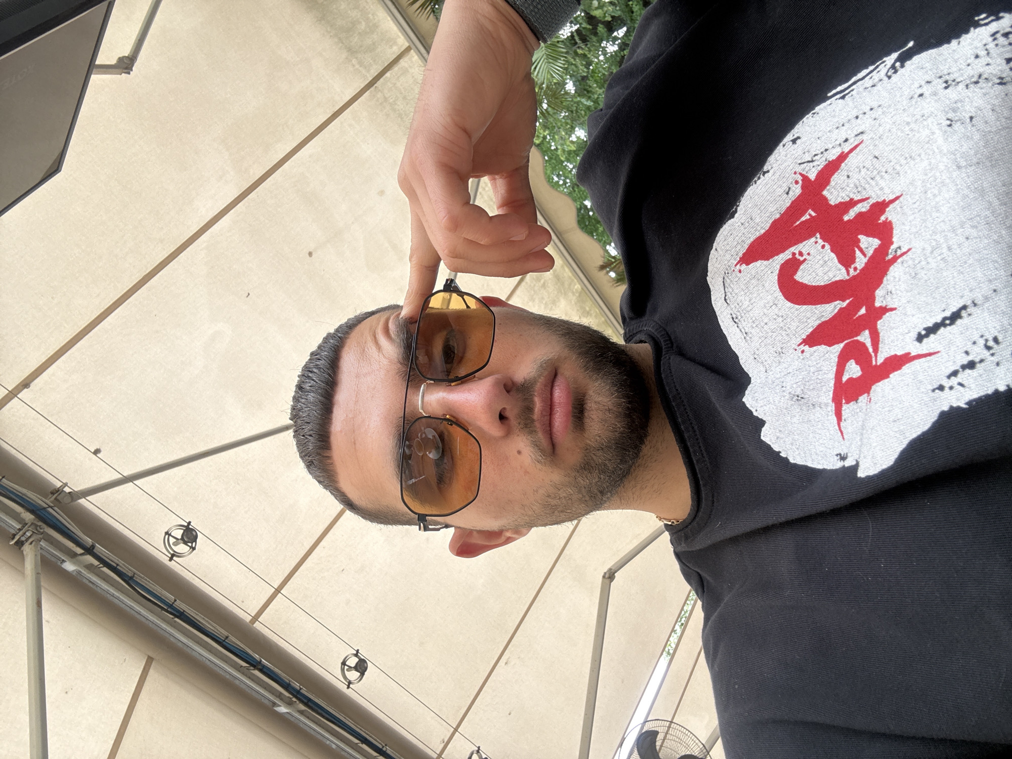 A man takes a selfie from a low angle, tilting the camera upward, outdoors on a bright day in Ruse, Bulgaria. He wears hexagonal-shaped gold-rimmed sunglasses and a black sweatshirt. The sweatshirt has a design on the chest consisting of a white blotch and red brushstroke-style text. His left hand rests on the edge of his sunglasses. The background shows a beige canvas awning supported by a metal frame, with electrical cables, pulleys, and glimpses of trees.