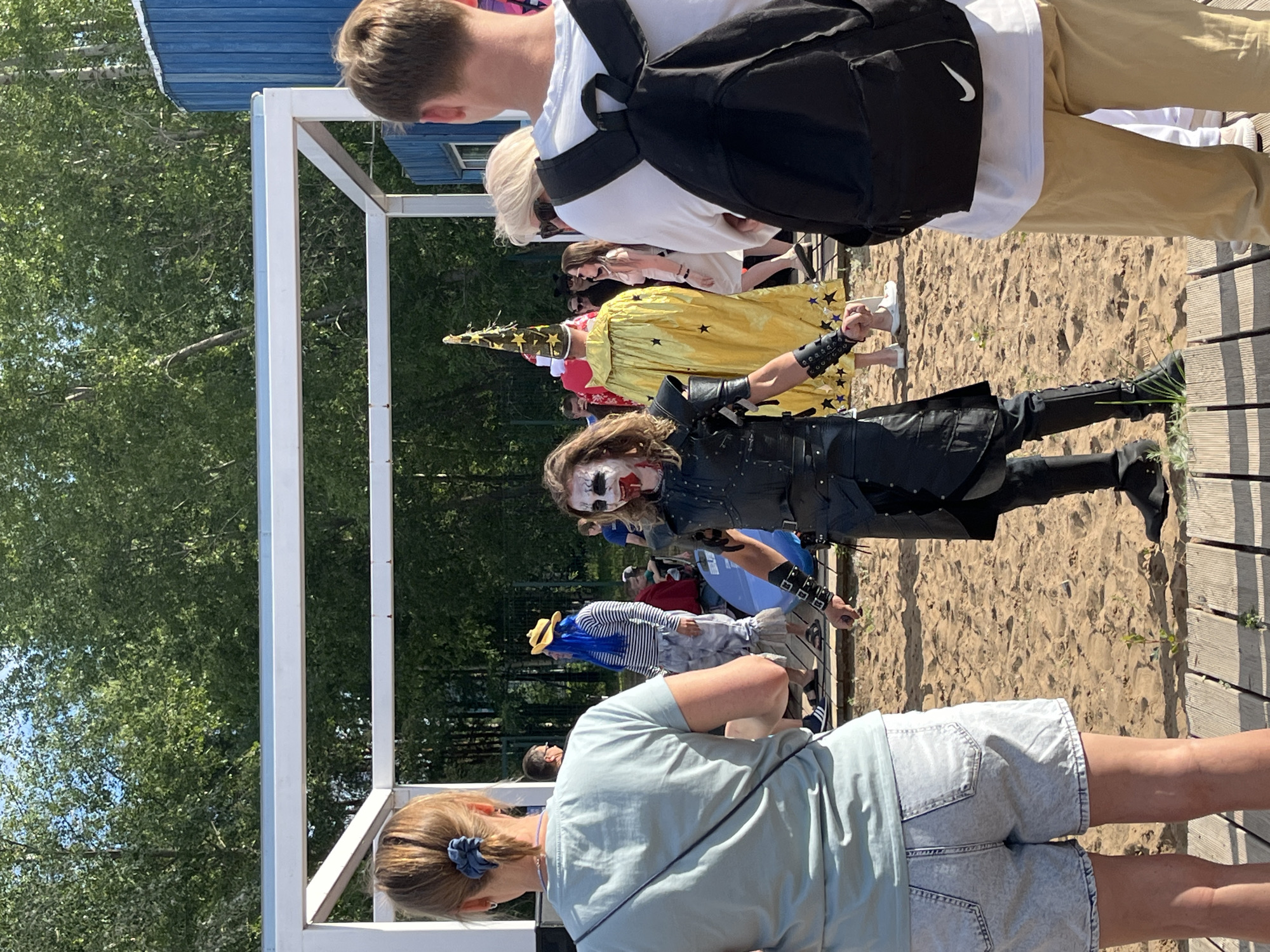 The image depicts an outdoor gathering, likely a festival or a performance, set against a backdrop of lush green trees and a sandy ground, possibly a beach or a park. The time of day appears to be sunny, with clear skies indicated by the bright illumination and sharp shadows.
The central subjects are individuals in costumes, suggesting a themed event or a costume parade. Prominently featured is a person dressed as a clown with exaggerated white face paint, dark eye makeup, and red lips. This individual wears a dark, possibly leather, outfit with intricate detailing and wristbands. Behind this person, another individual is dressed in a vibrant yellow costume adorned with black stars, topped with a tall, pointed hat that also has star embellishments. Further back, a person with bright blue hair and a striped outfit can be seen. In the foreground, the back of a person wearing a light blue t-shirt and denim shorts is visible, looking towards the costumed individuals. Another person in the foreground has their back to the camera, wearing a white t-shirt and a black backpack, suggesting they are also attending the event.
The scene unfolds on a wooden boardwalk or platform adjacent to the sandy area. White structural elements, possibly part of a temporary stage or canopy, frame the scene. The presence of multiple people in distinct costumes implies a celebratory or entertainment-focused event. The overall mood seems lively and casual, fitting for a daytime outdoor gathering. No discernible text is visible in the image.